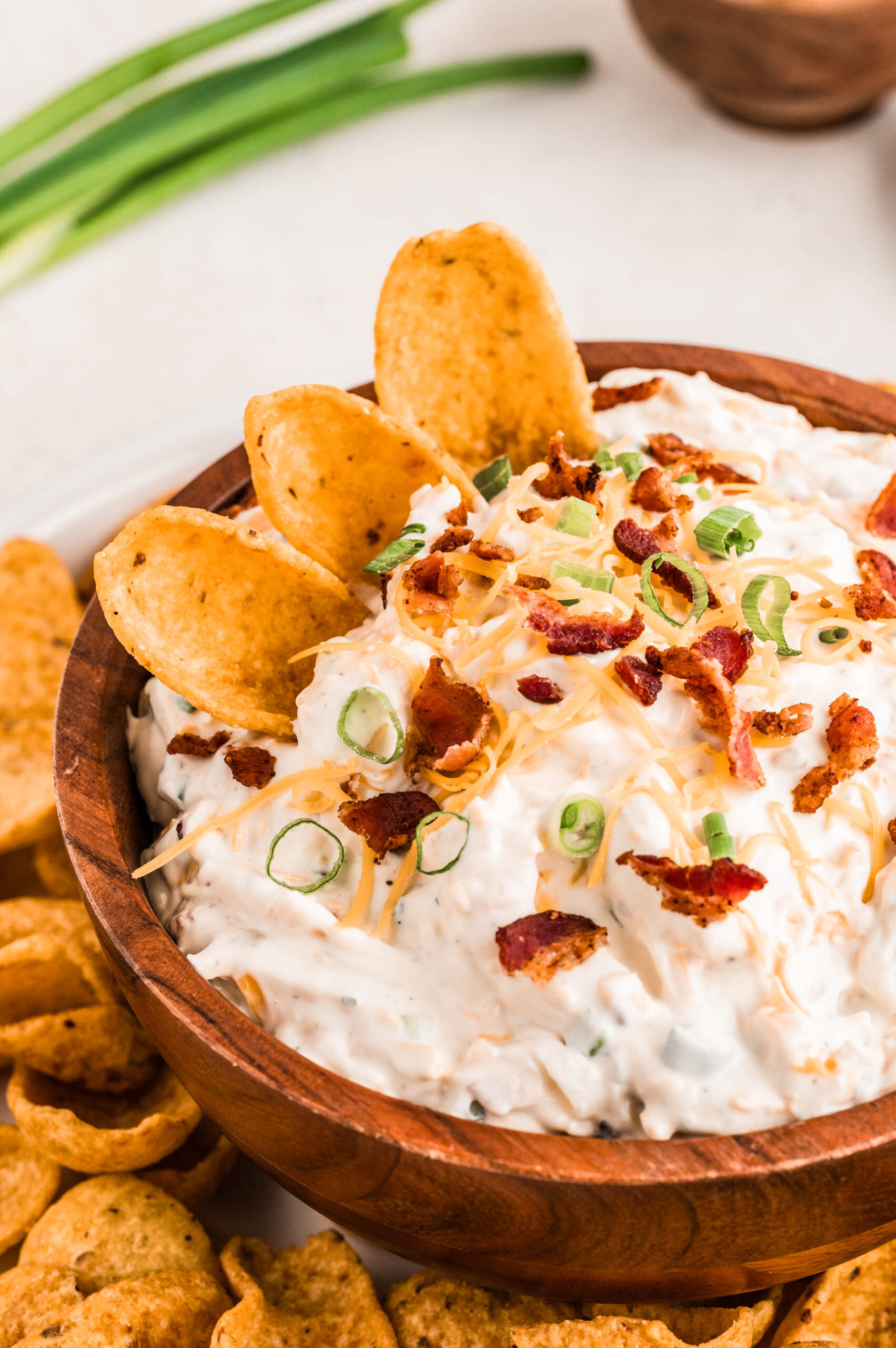 Close-up of creamy bacon ranch cheddar dip in a wooden bowl, topped with crispy bacon pieces, shredded cheddar cheese, and sliced green onions, with corn chips dipped into the top and scattered around the bowl.