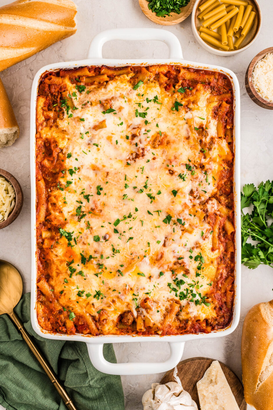 A large white casserole dish filled with golden, bubbly no boil baked ziti topped with melted mozzarella and sprinkled with fresh parsley. The dish is surrounded by ingredients including uncooked ziti, grated parmesan, fresh herbs, and sliced baguette on a light countertop.