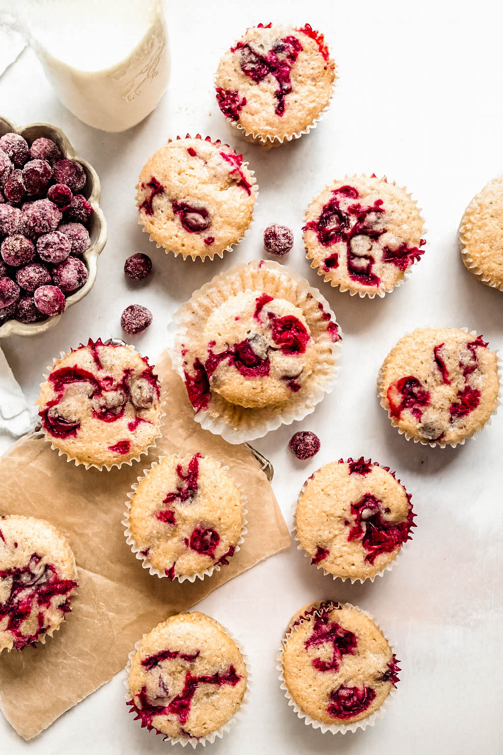 Overhead view of cranberry orange muffins scattered across a white marble surface, showing golden tops studded with vibrant red cranberries. A bowl of sugared cranberries and a small milk bottle sit to the side, adding a cozy, homemade feel to the scene.