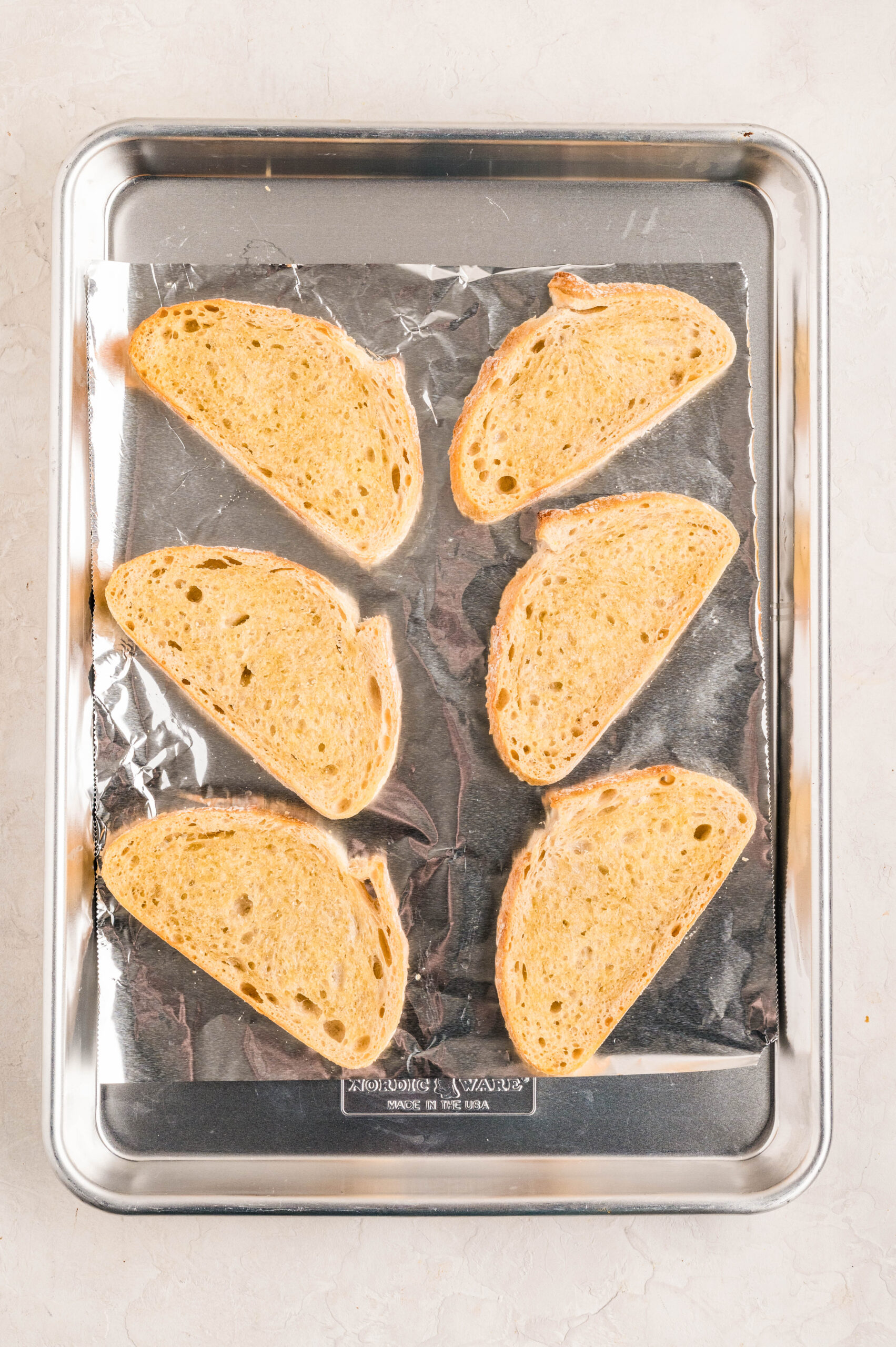 Six slices of crusty bread arranged on a foil-lined baking sheet, lightly toasted and ready to be topped for making Smoked Salmon Bruschetta