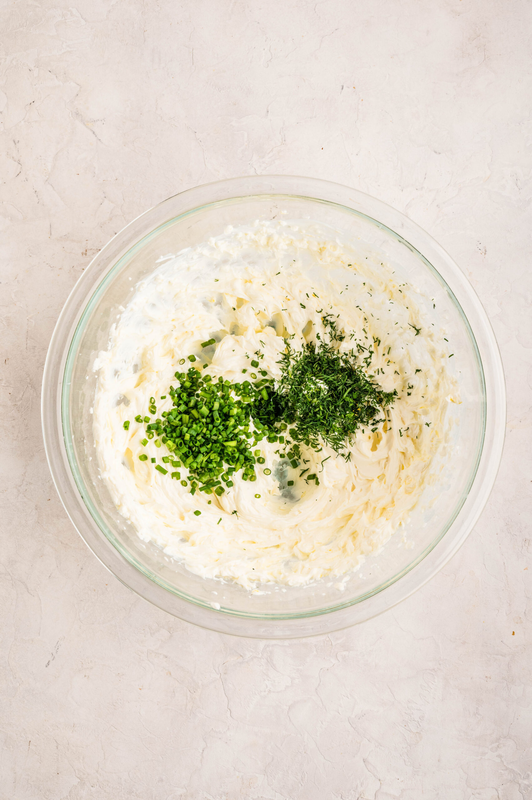 Glass bowl filled with whipped cream cheese and crème fraîche, topped with freshly chopped chives and dill, ready to be mixed.