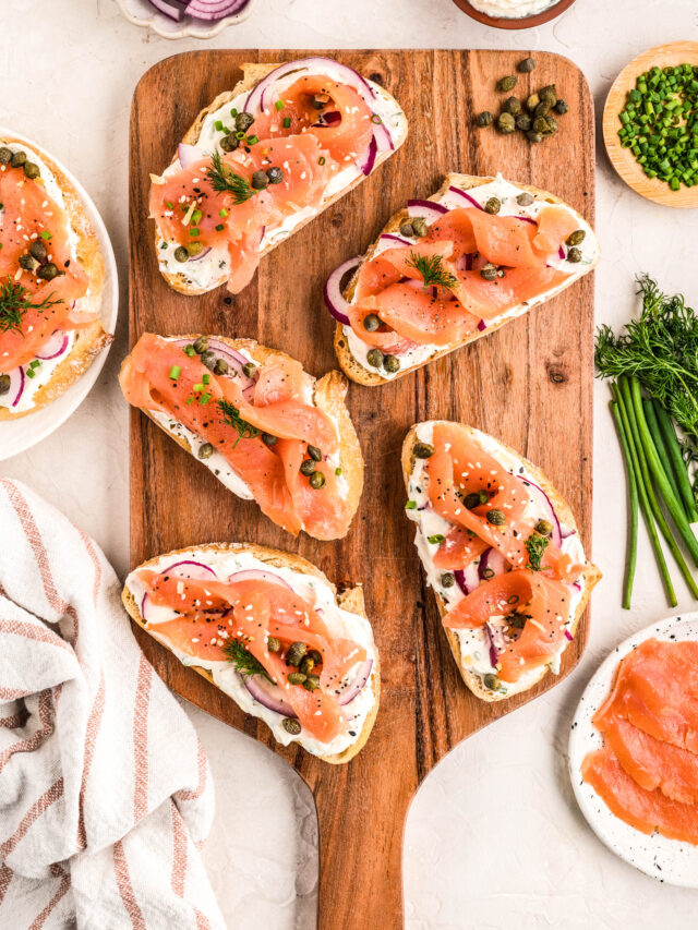 Smoked Salmon Bruschetta arranged on a wooden serving board, topped with a creamy lemon-herb spread, thin red onion slices, capers, fresh dill, and chives. Additional ingredients like sliced red onions, chives, dill, and smoked salmon surround the board for a fresh, vibrant presentation