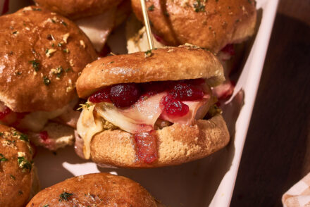 Close-up of baked Thanksgiving turkey leftover sliders in a white baking dish, with golden toasted rolls brushed with garlic herb butter, melted provolone cheese, stuffing, and cranberry sauce visible inside one slider secured with a toothpick.