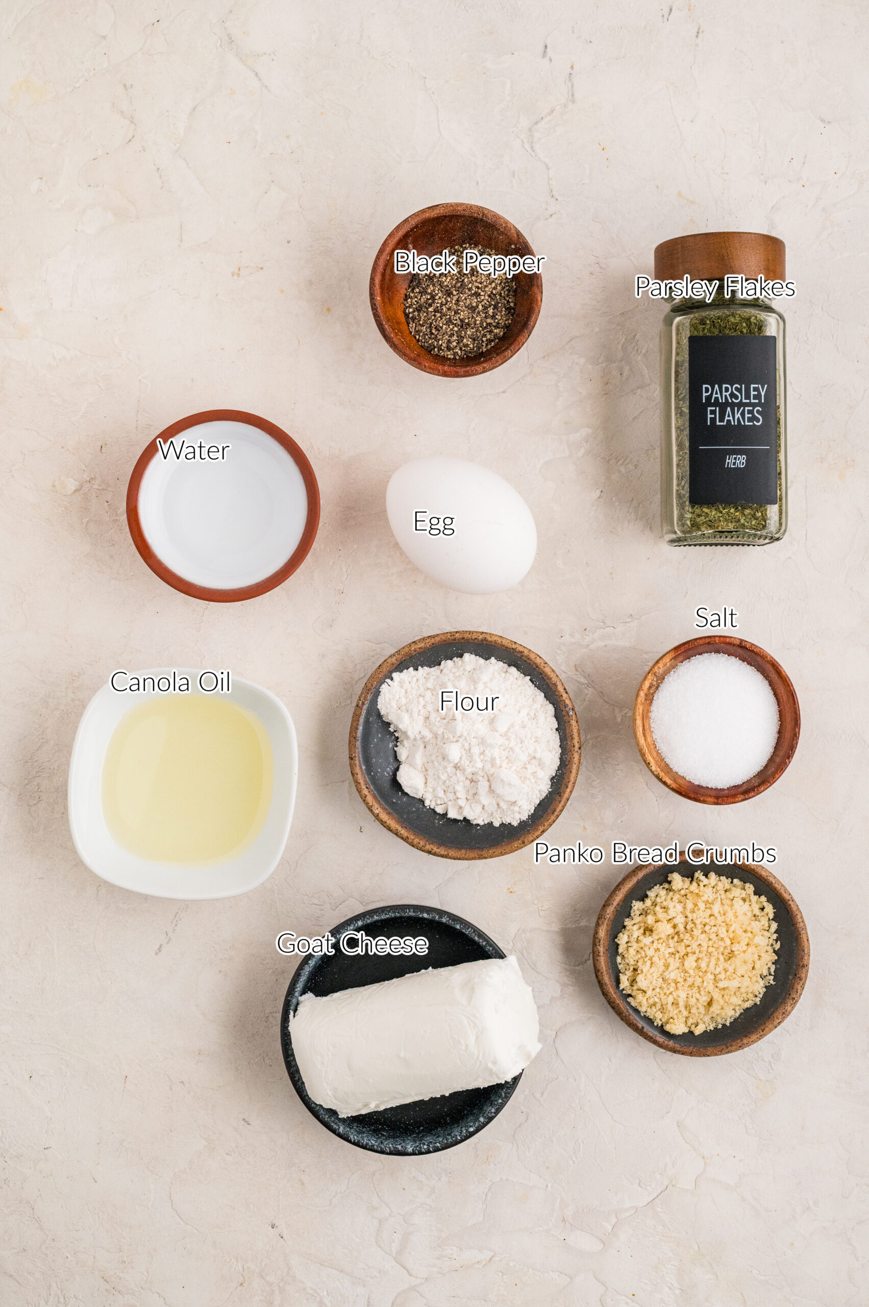 Overhead image of ingredients for fried goat cheese balls arranged neatly on a light surface, including a log of goat cheese, flour, panko breadcrumbs, salt, black pepper, parsley flakes, an egg, water, and canola oil in small bowls, each labeled for reference.