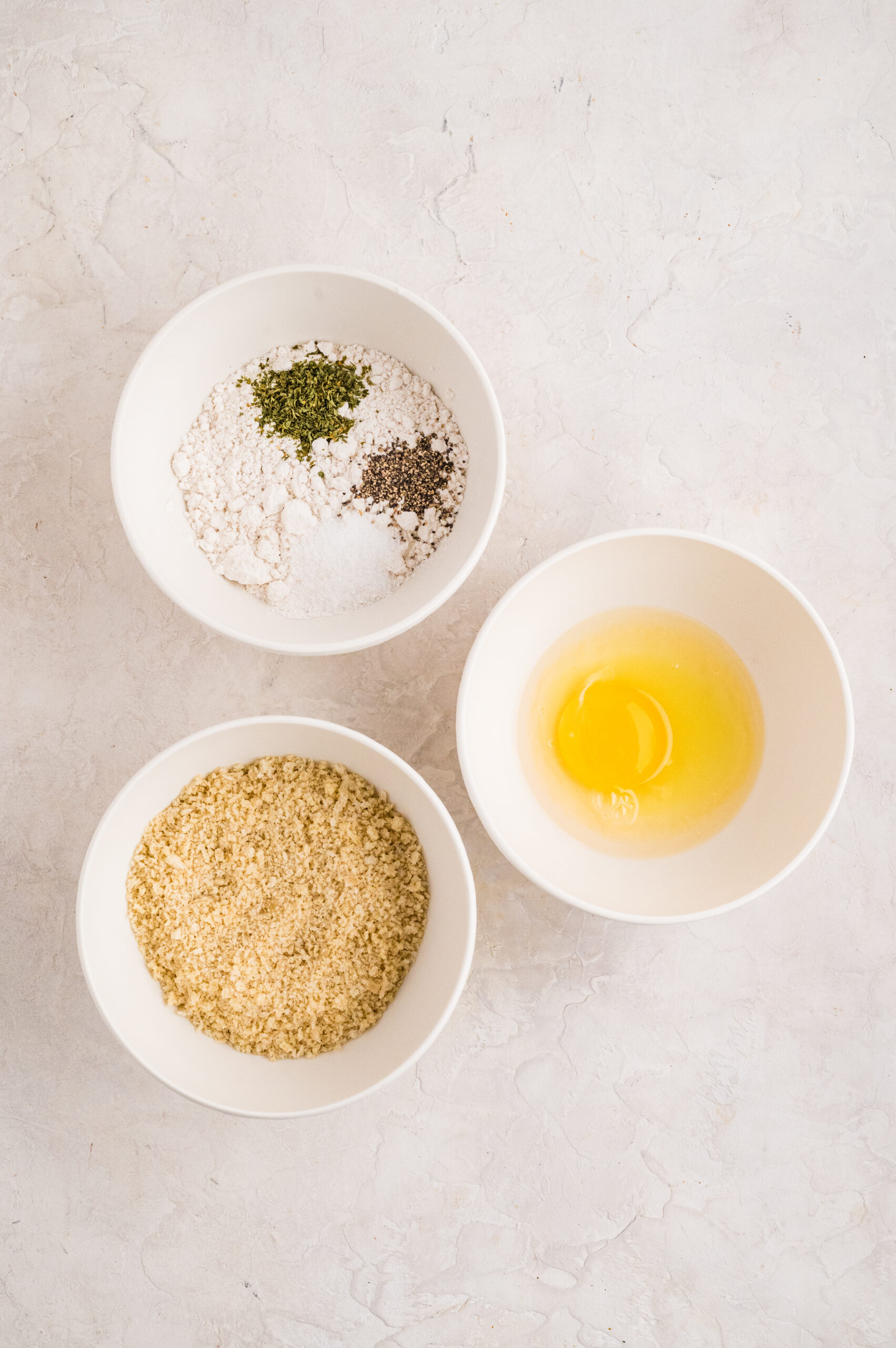 Overhead view of three bowls showing the dredging station for fried goat cheese balls: one bowl with flour mixed with parsley, salt, and black pepper, a second bowl with a cracked egg for the egg wash, and a third filled with panko breadcrumbs, all arranged on a light textured surface.