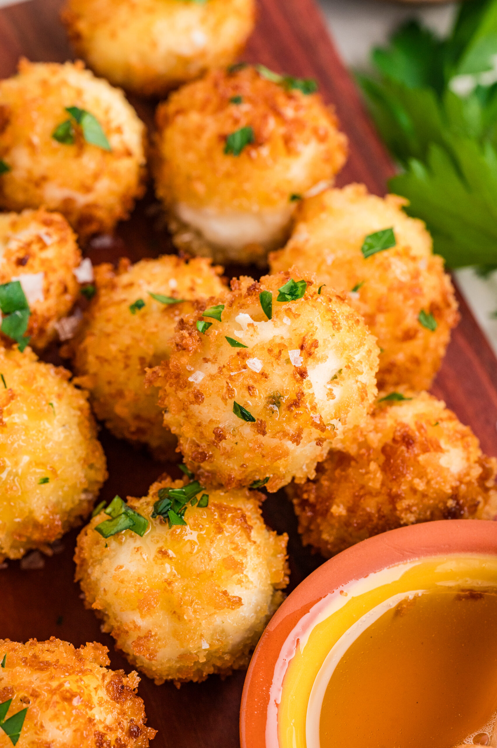Close-up shot of crispy fried goat cheese balls sprinkled with fresh parsley and flaky sea salt, showing their golden panko coating beside a small bowl of honey for dipping.