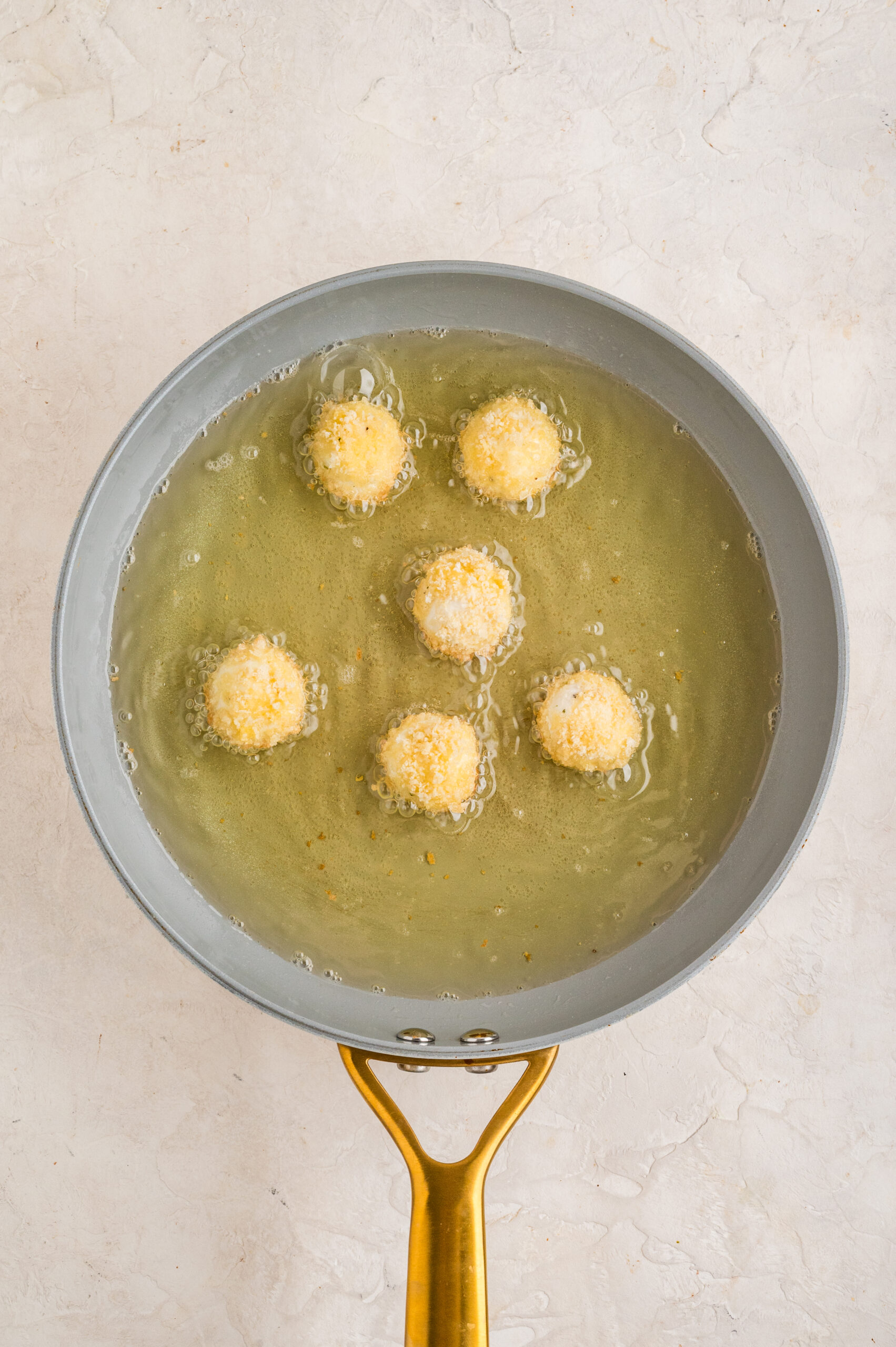 Overhead view of a skillet filled with hot oil as several coated goat cheese balls fry, bubbling gently as they cook and begin to turn golden, showing the first stage of frying for fried goat cheese balls.