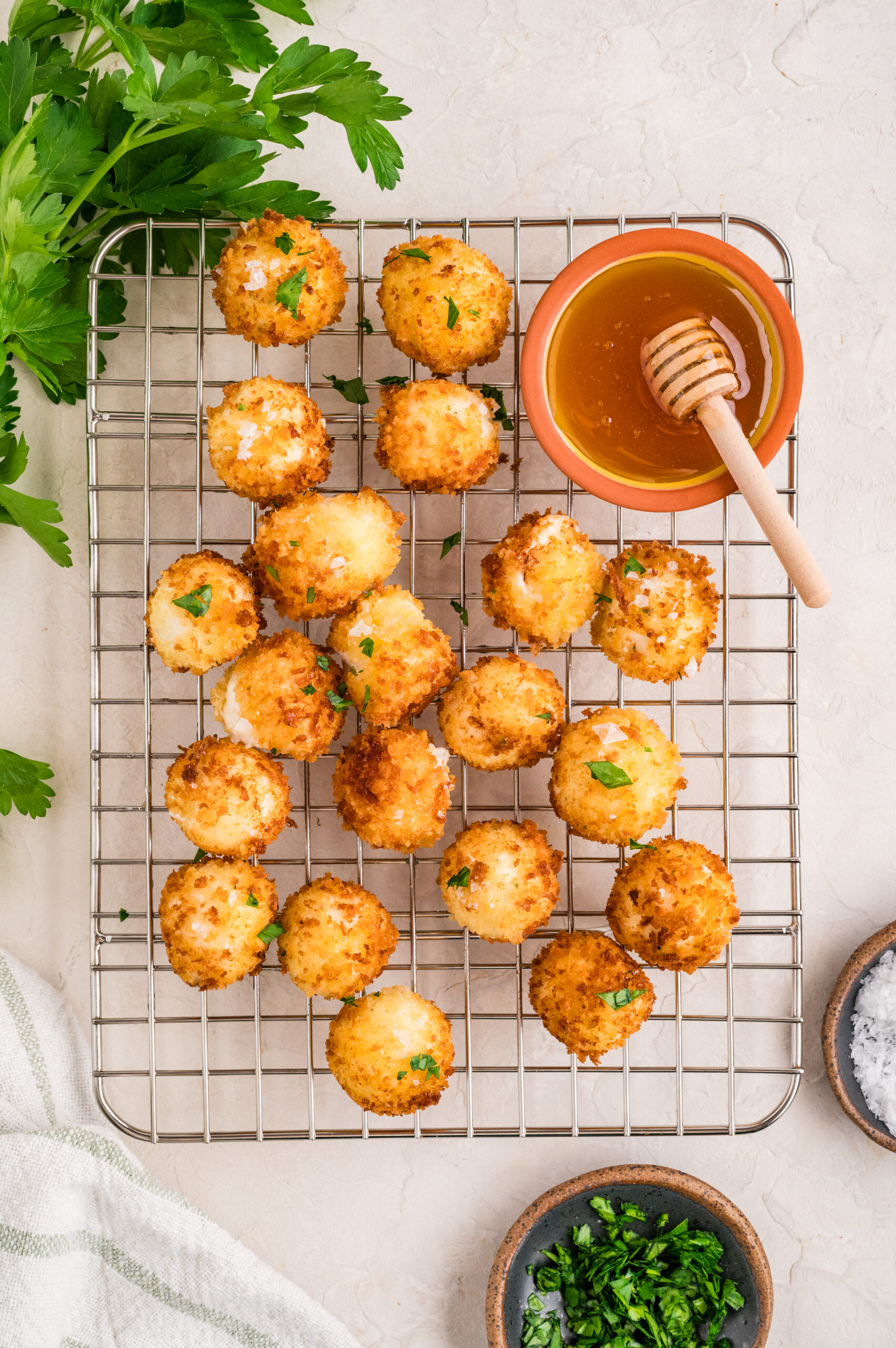 Overhead image of golden fried goat cheese balls resting on a cooling rack, garnished with fresh parsley and flaky sea salt, served alongside a small bowl of honey with a honey dipper, creating a bright and appetizing presentation.