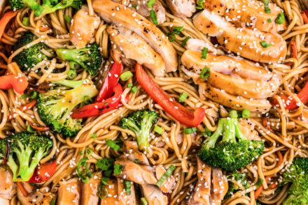 A large white skillet filled with Chicken Teriyaki Bowls featuring sliced teriyaki-glazed chicken thighs, lo mein noodles, bright broccoli florets, red bell pepper strips, and shredded carrots, all tossed in a glossy homemade teriyaki sauce and topped with sesame seeds and green onions.
