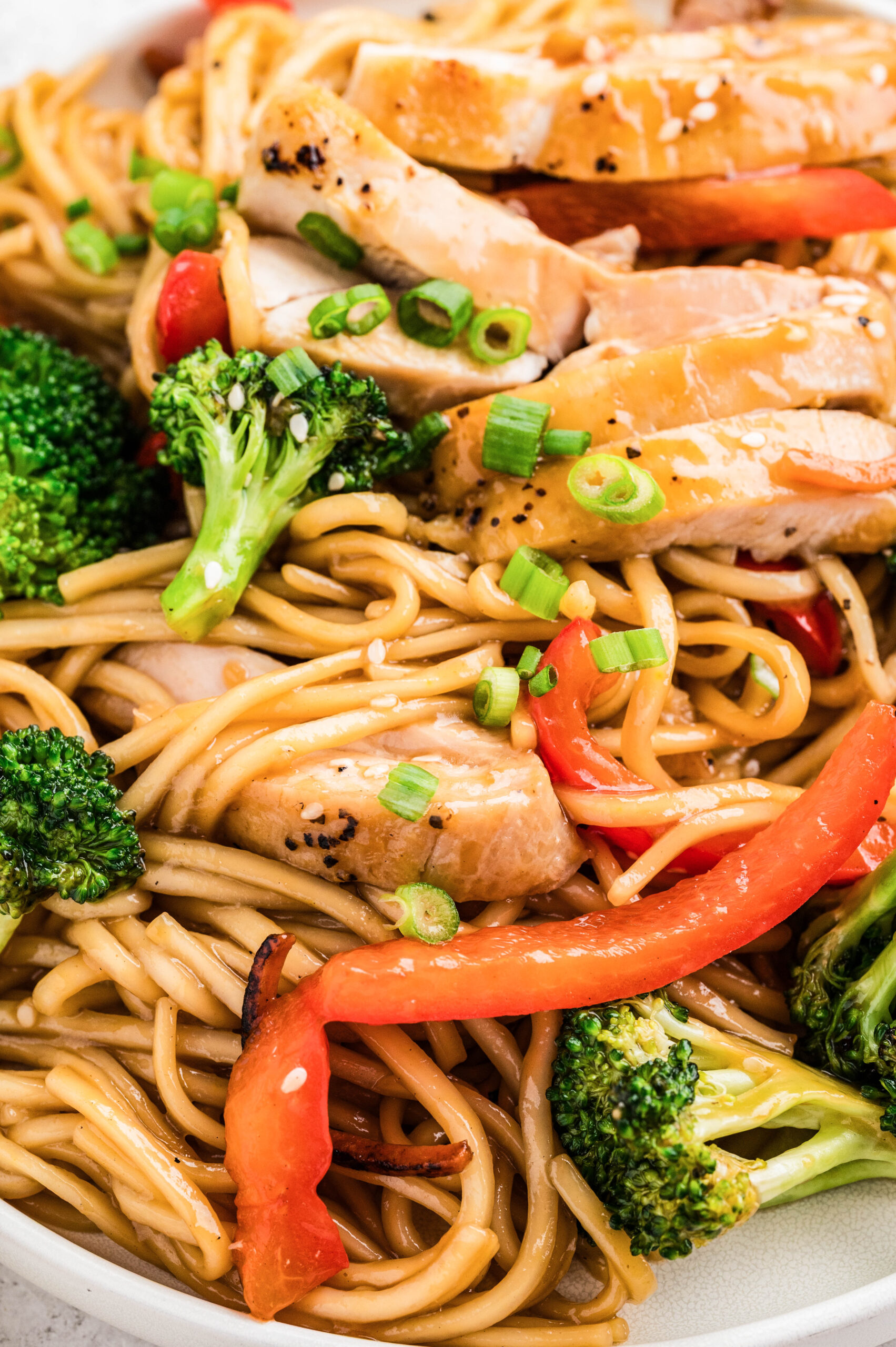 A close-up of Chicken Teriyaki Bowls showing glossy lo mein noodles, sliced teriyaki chicken thighs, bright red bell pepper strips, crisp broccoli florets, and a sprinkle of green onions and sesame seeds.