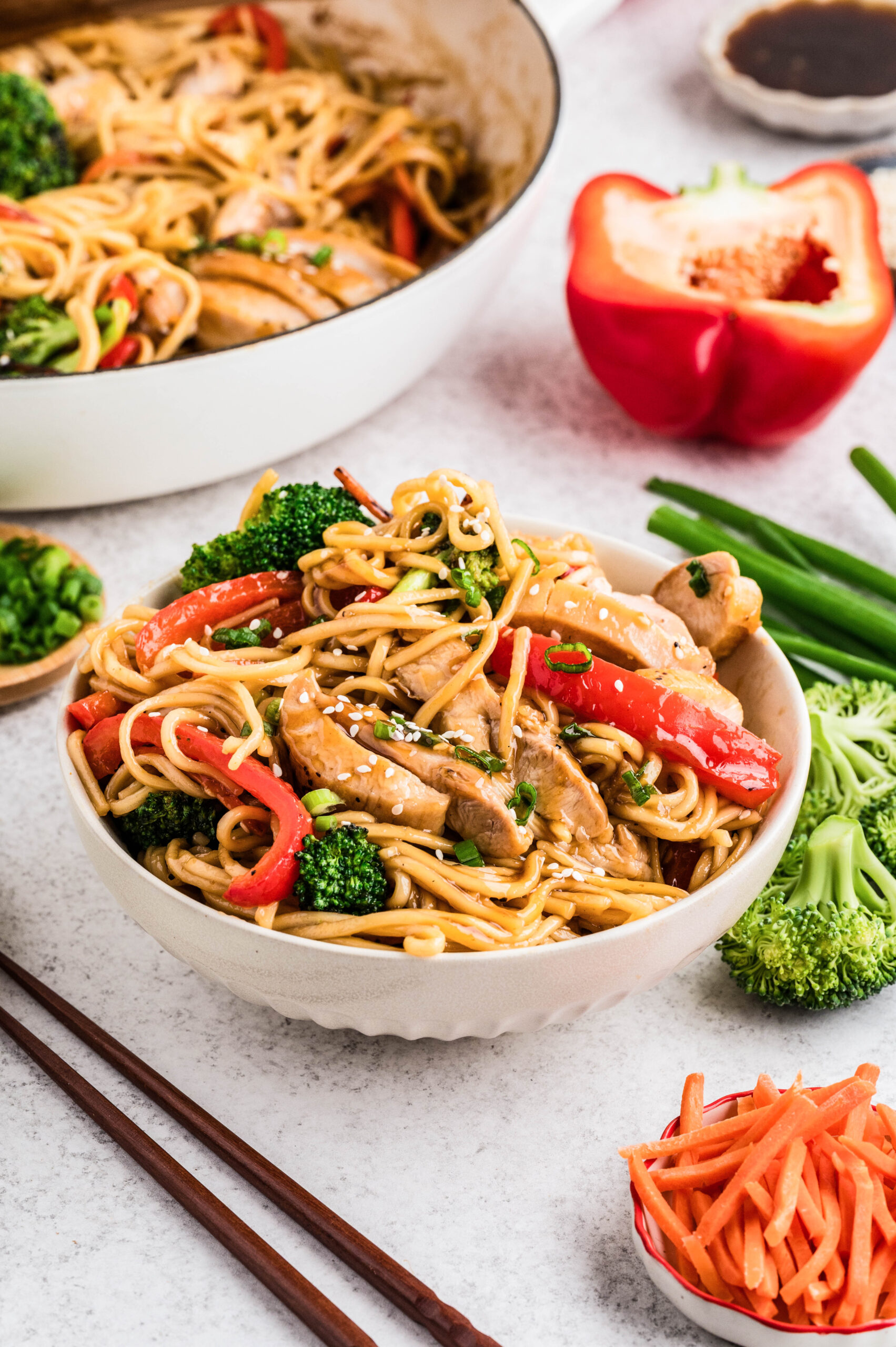 A bowl of Chicken Teriyaki Bowls filled with saucy lo mein noodles, sliced teriyaki-glazed chicken, broccoli florets, and red bell pepper strips, topped with sesame seeds and green onions. Fresh vegetables, shredded carrots, and a pan of noodles sit in the background.