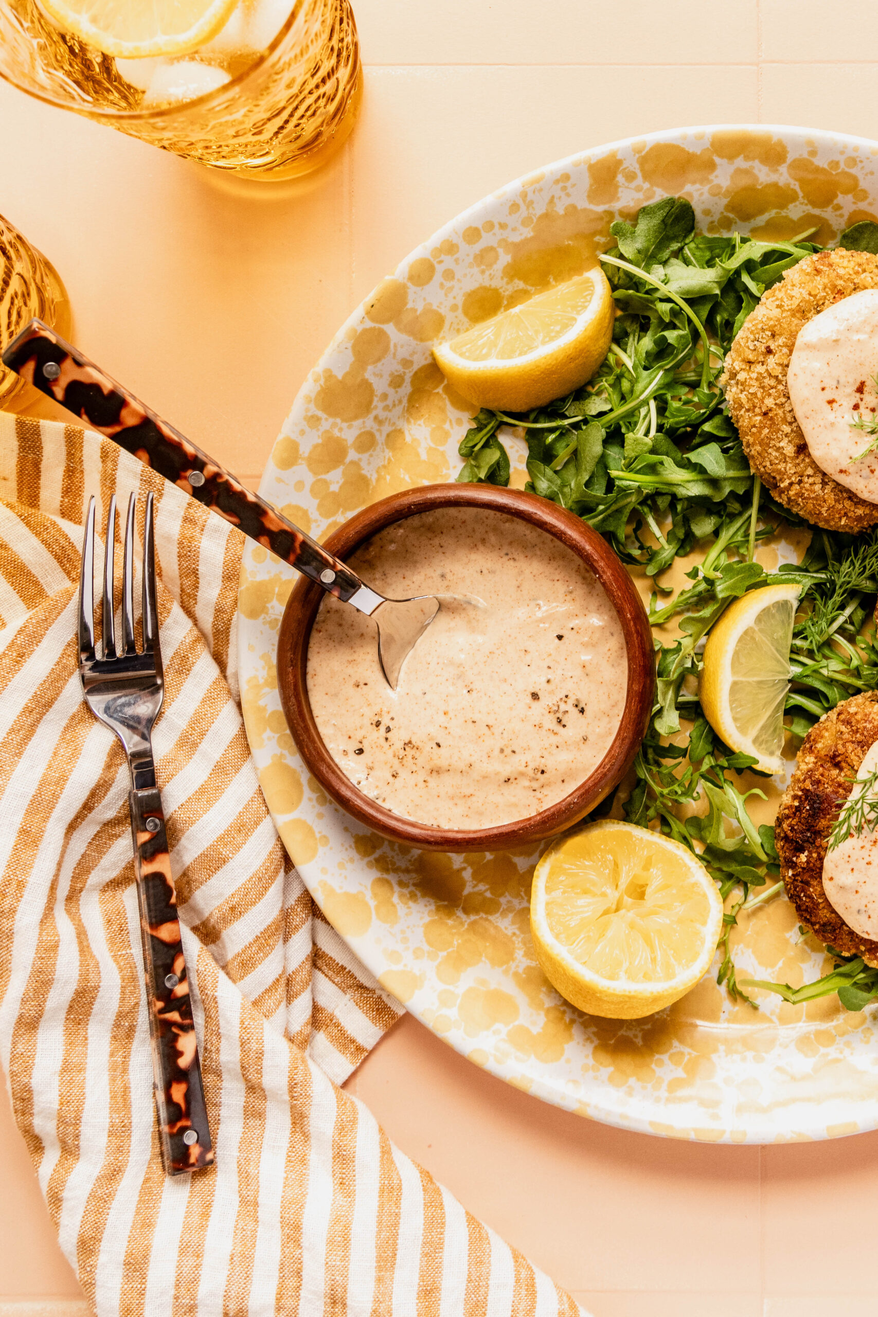 Overhead view of a creamy homemade Remoulade Sauce in a small bowl with a spoon, surrounded by crispy patties, fresh greens, and lemon wedges on a warm-toned plate with striped napkin and glassware.