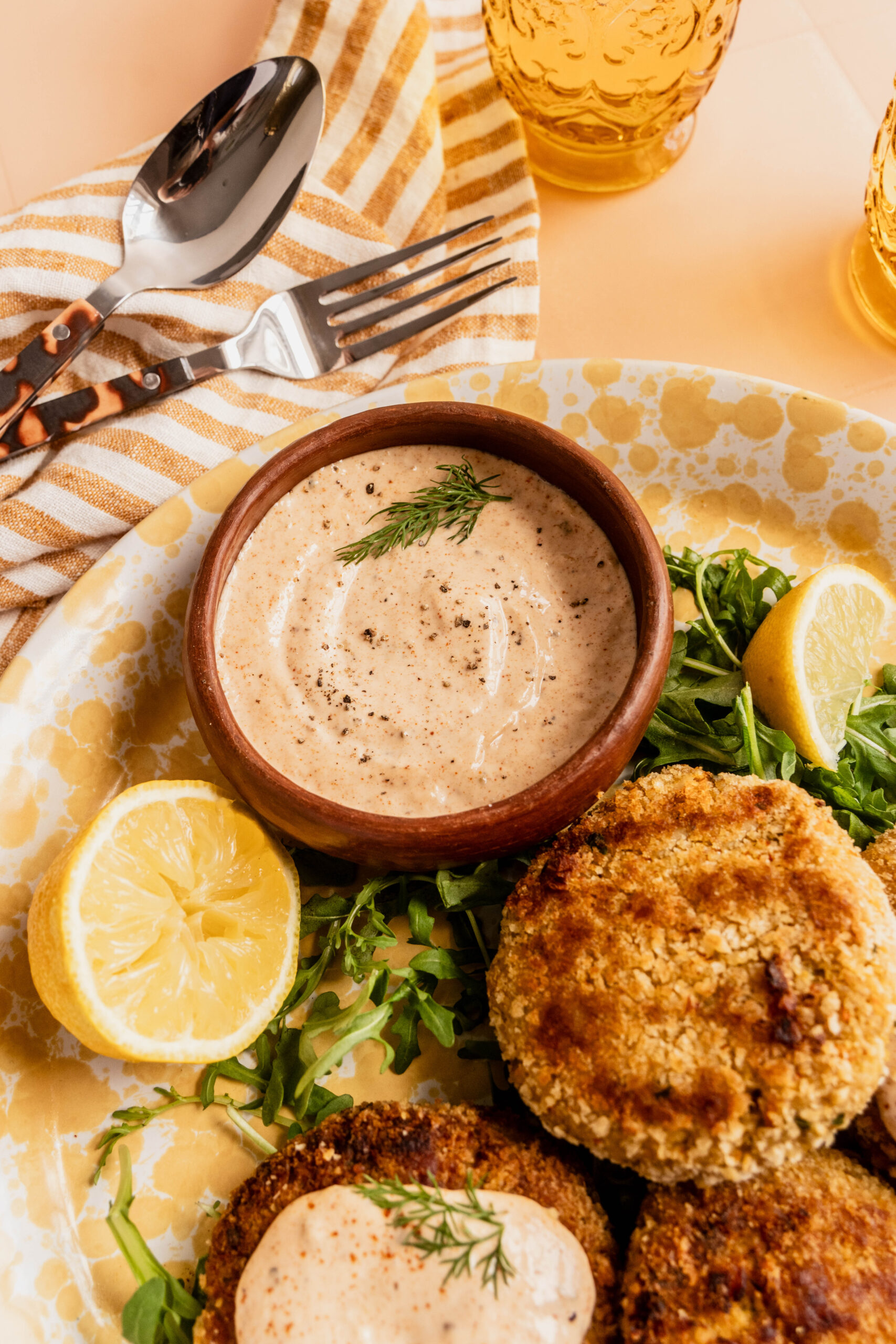 Close-up of a bowl of creamy homemade Remoulade Sauce sprinkled with black pepper and herbs, served with crispy golden patties, fresh arugula, and lemon wedges on a patterned plate.