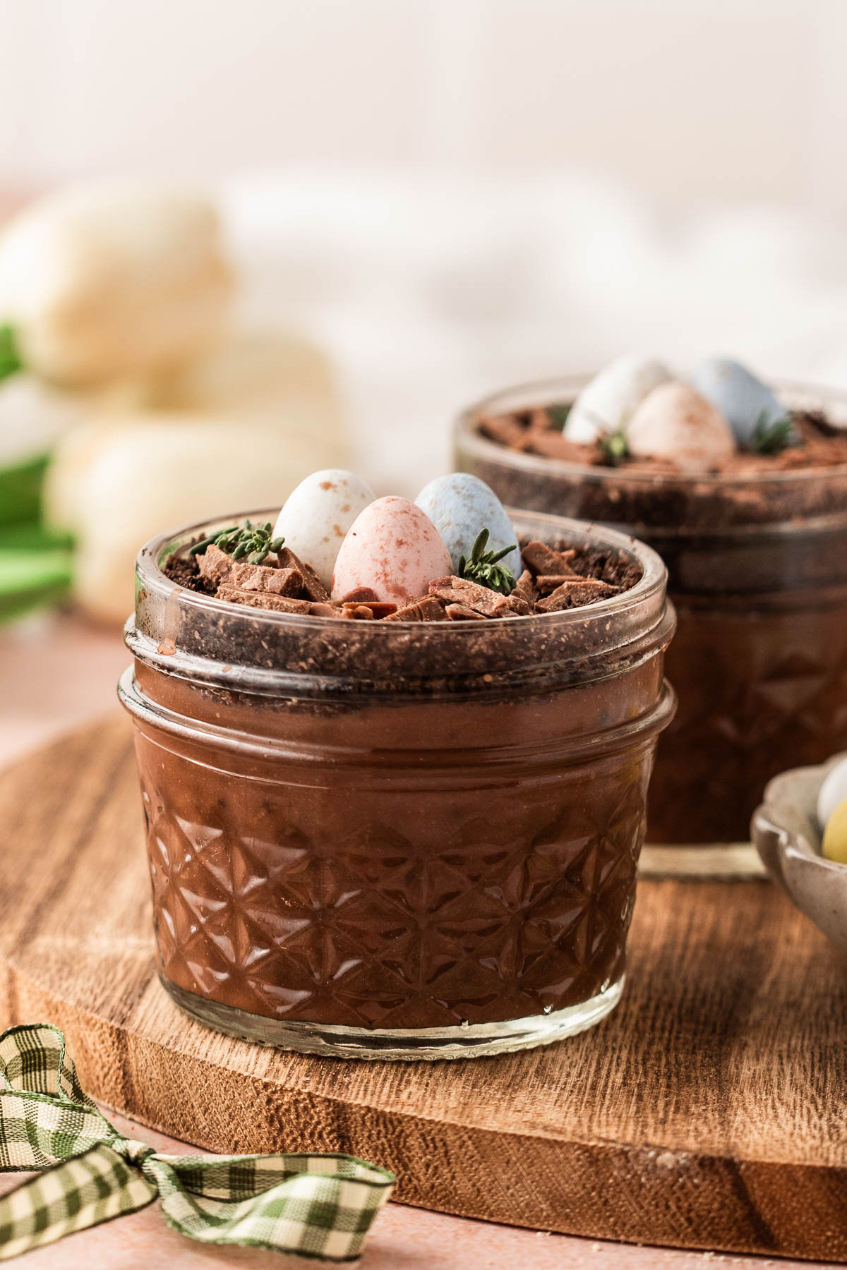 Easter dirt cups served in clear glass jars with layers of chocolate pudding and Oreo crumb “dirt,” topped with chocolate nests, speckled pastel mini eggs, and fresh thyme on a wooden board.