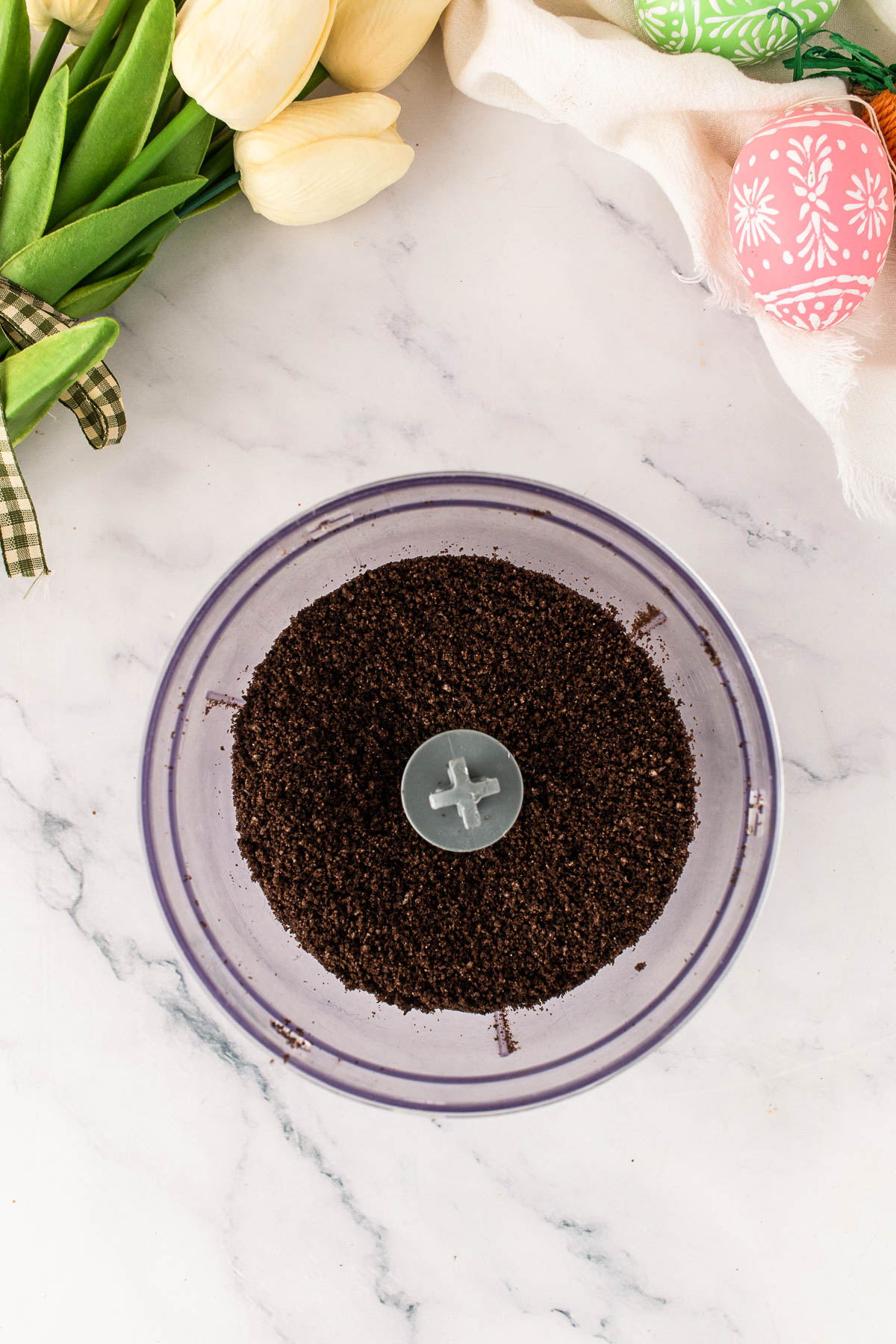 Finely crushed Oreo cookies in a food processor, prepared as the dirt layer for Easter dirt cups, styled on a white marble surface with spring tulips and pastel Easter eggs.