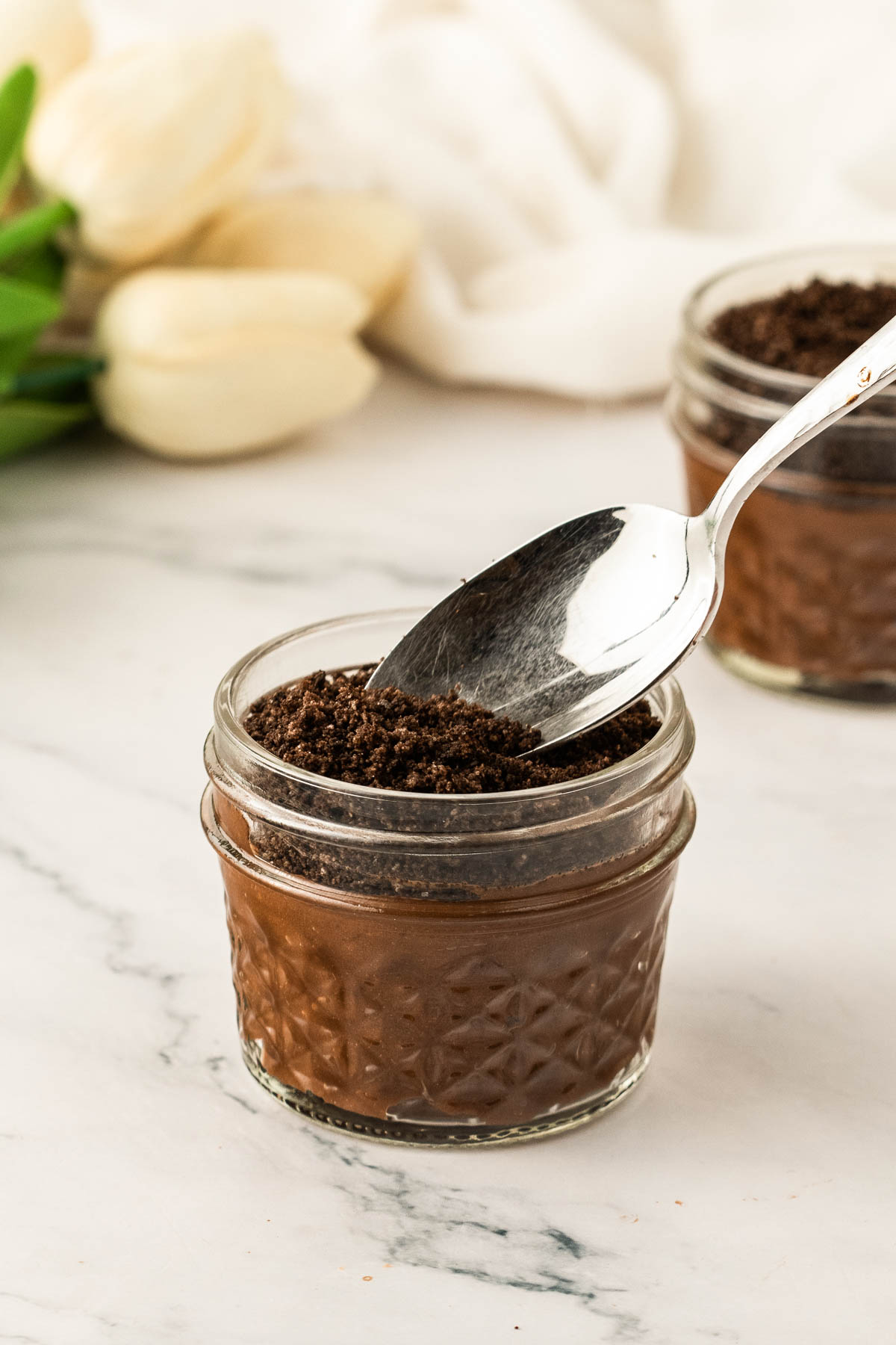Layering Oreo crumb “dirt” over chocolate pudding in a glass jar while assembling Easter dirt cups, with a spoon gently smoothing the top on a white marble surface.