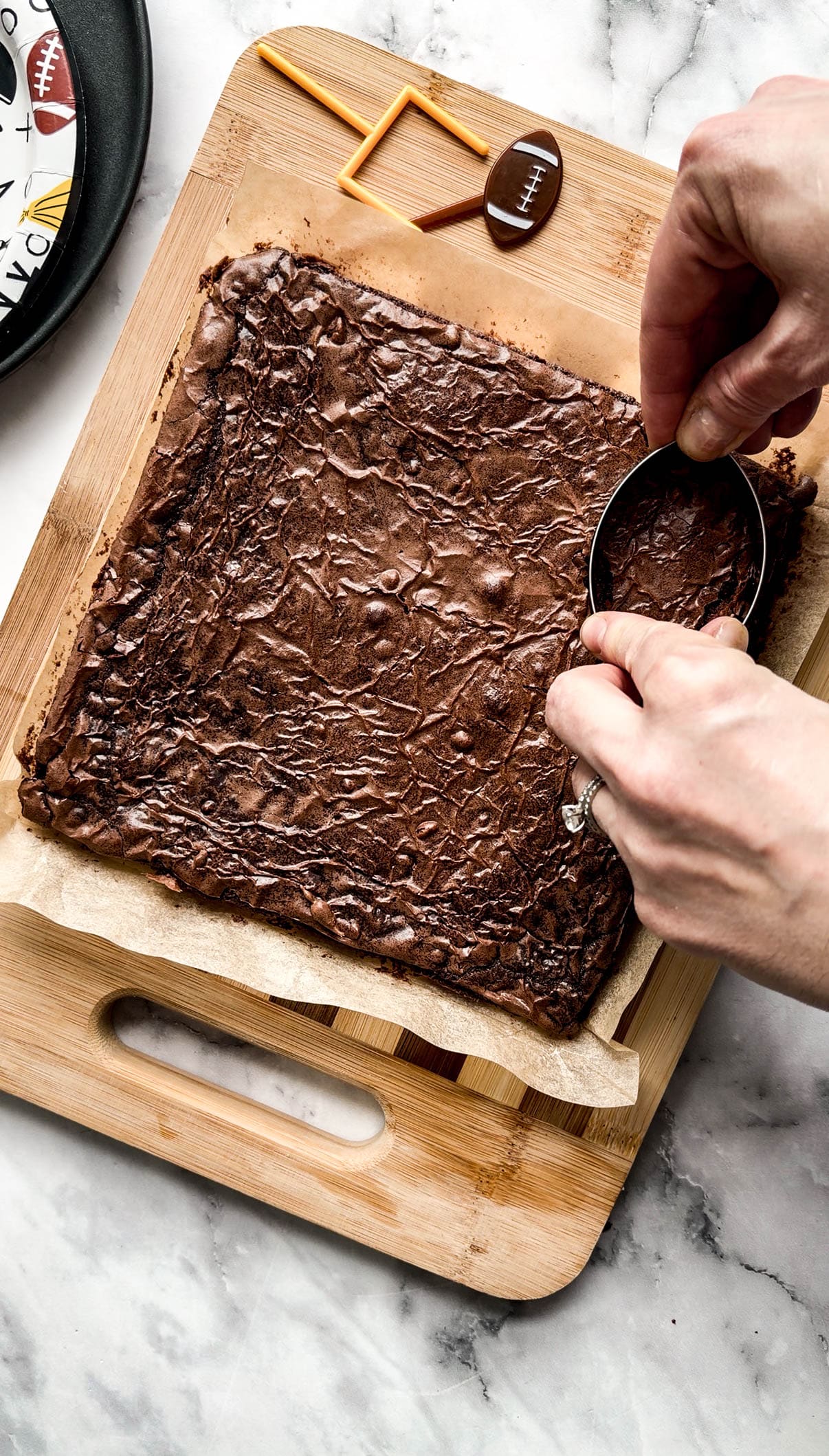 Overhead view of a fudgy pan of football brownies being cut into football shapes with a cookie cutter on a wooden cutting board