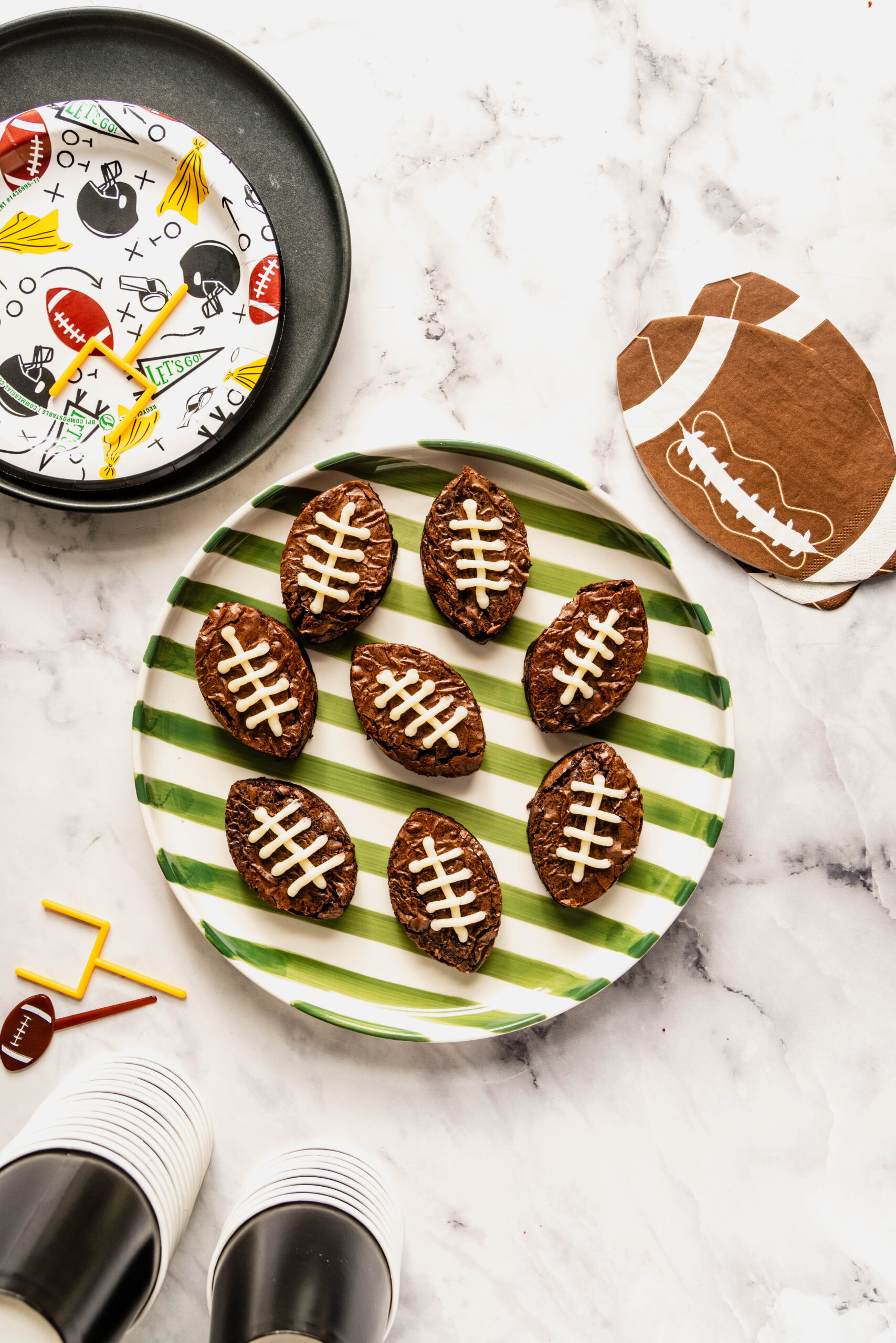 Overhead view of fudgy football brownies decorated with white buttercream laces, arranged on a green-striped plate on a white marble background with football-themed party plates, napkins, and cups.