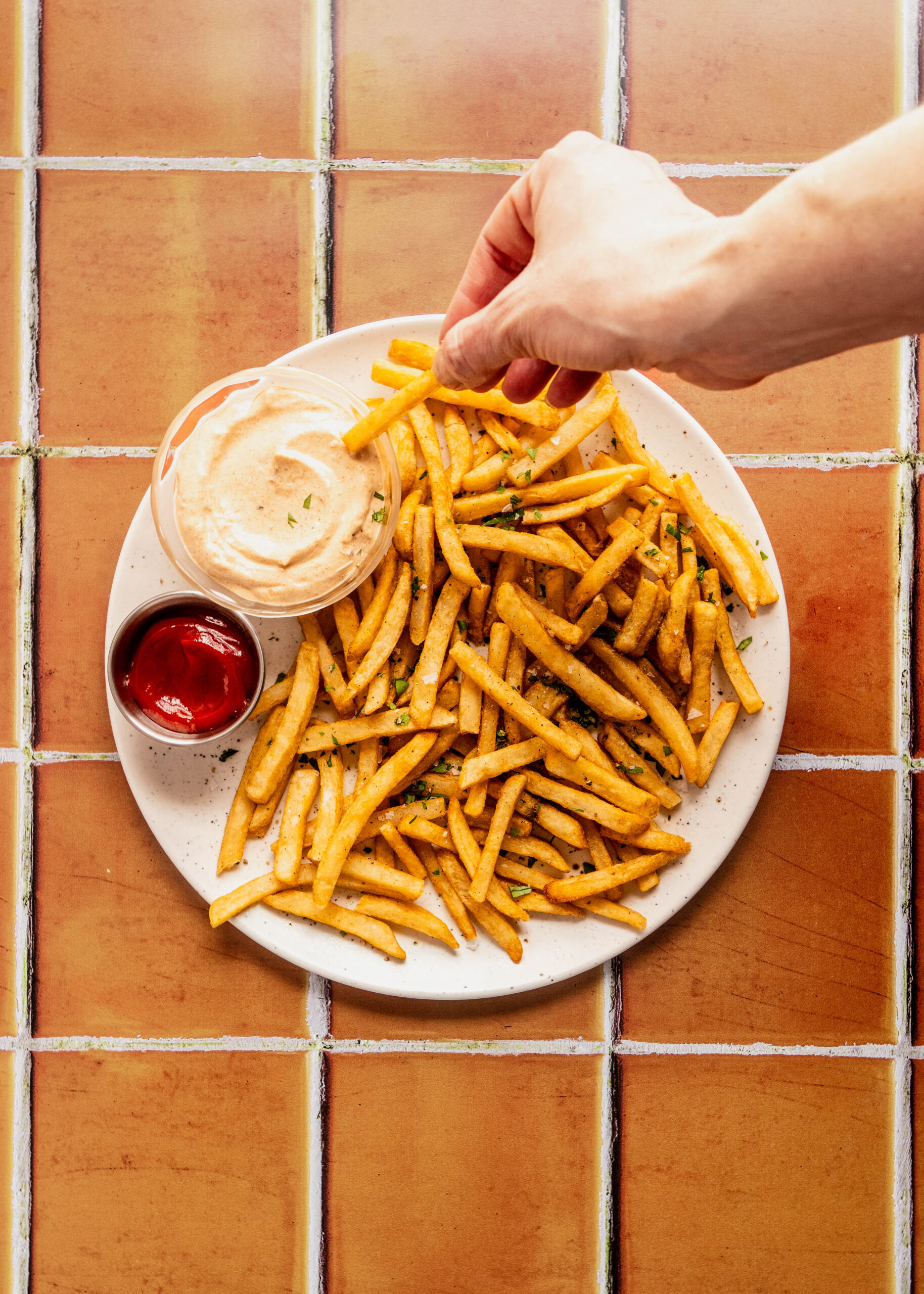 Overhead view of crispy French fries on a plate with a hand dipping a fry into creamy Remoulade Sauce, served alongside ketchup on a warm tiled background.