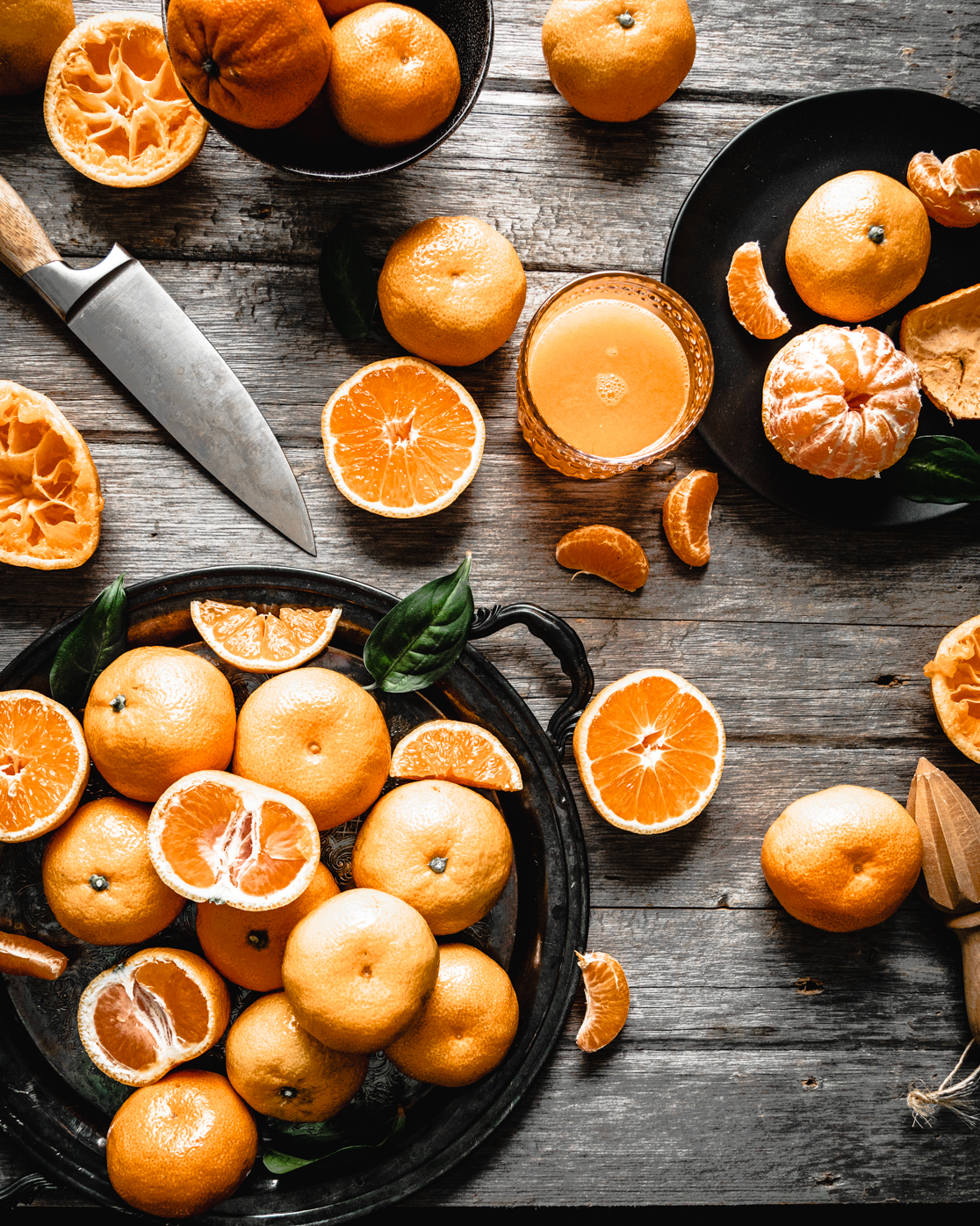 Rustic overhead scene of fresh oranges, halved and peeled, with a knife and a glass of freshly squeezed orange juice on a wooden table, showing citrus prep for an orange curd recipe.