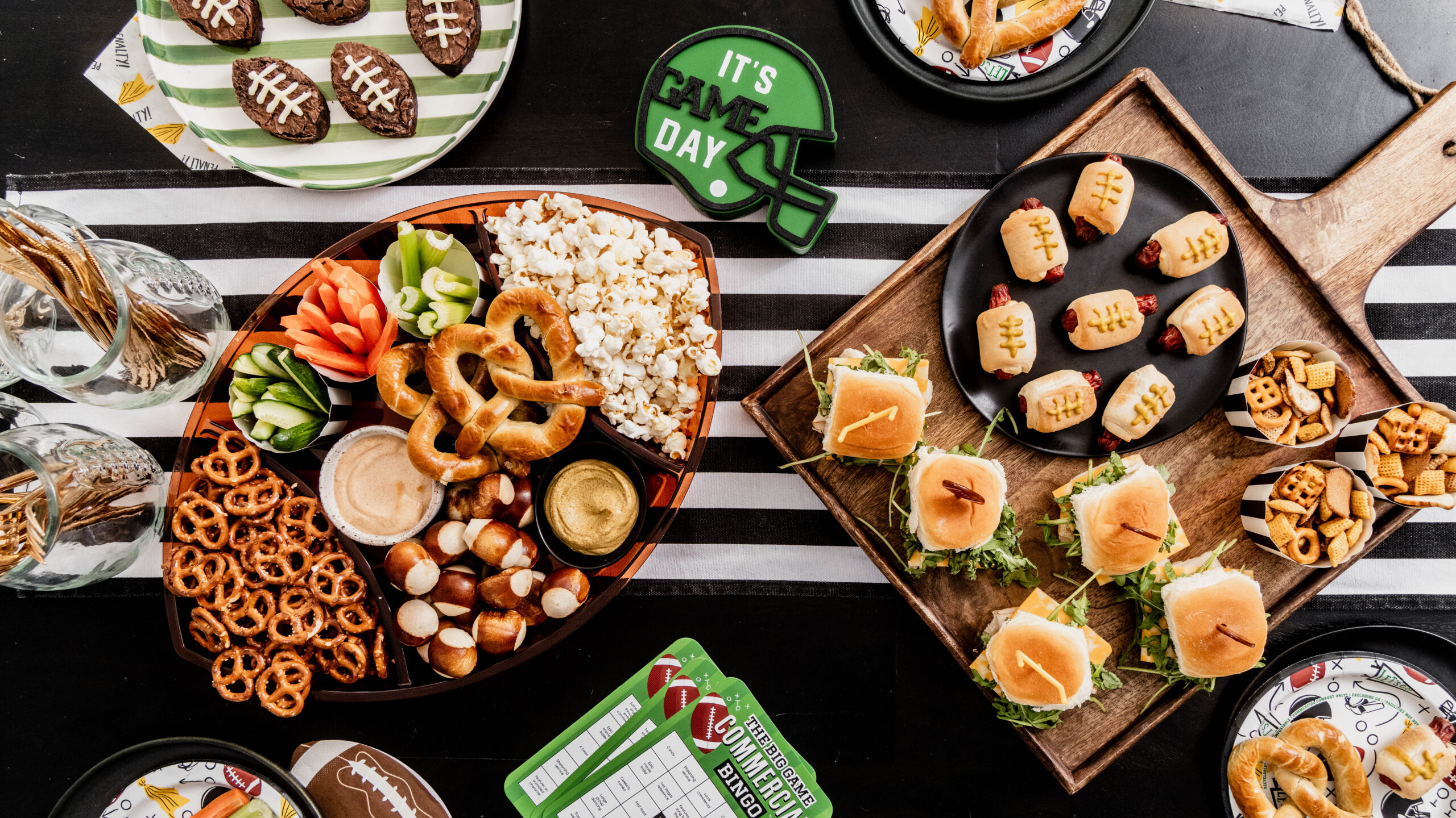 Overhead game day tablescape featuring football brownies with white buttercream laces alongside sliders, pretzels, popcorn, veggie snacks, and football-themed plates and decor on a black and white striped table.