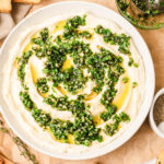Overhead view of whipped brie spread in a white bowl, topped with chopped fresh herbs and olive oil, surrounded by crostini, crackers, and sliced apples on a light marble surface.