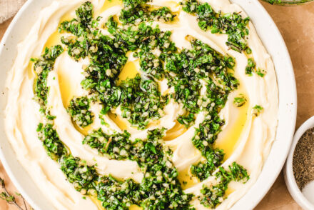 Overhead view of whipped brie spread in a white bowl, topped with chopped fresh herbs and olive oil, surrounded by crostini, crackers, and sliced apples on a light marble surface.