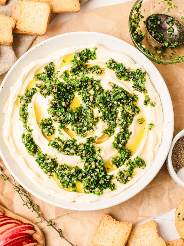 Overhead view of whipped brie spread in a white bowl, topped with chopped fresh herbs and olive oil, surrounded by crostini, crackers, and sliced apples on a light marble surface.