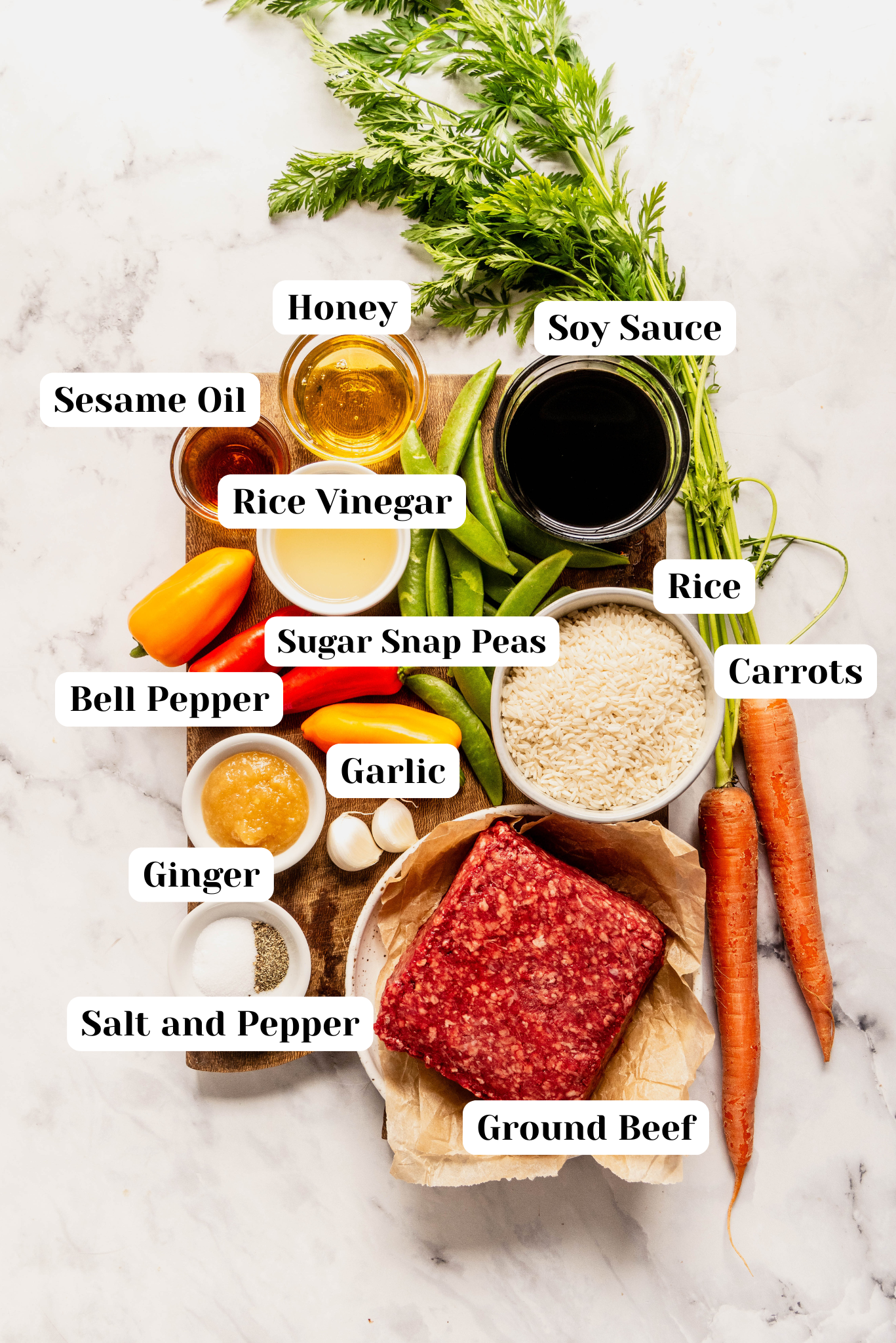 Overhead ingredient shot for a Korean beef rice bowl featuring ground beef, white rice, soy sauce, honey, sesame oil, rice vinegar, garlic, ginger, sugar snap peas, bell peppers, carrots, and salt and pepper arranged on a wooden board over a white marble background, with each ingredient labeled.