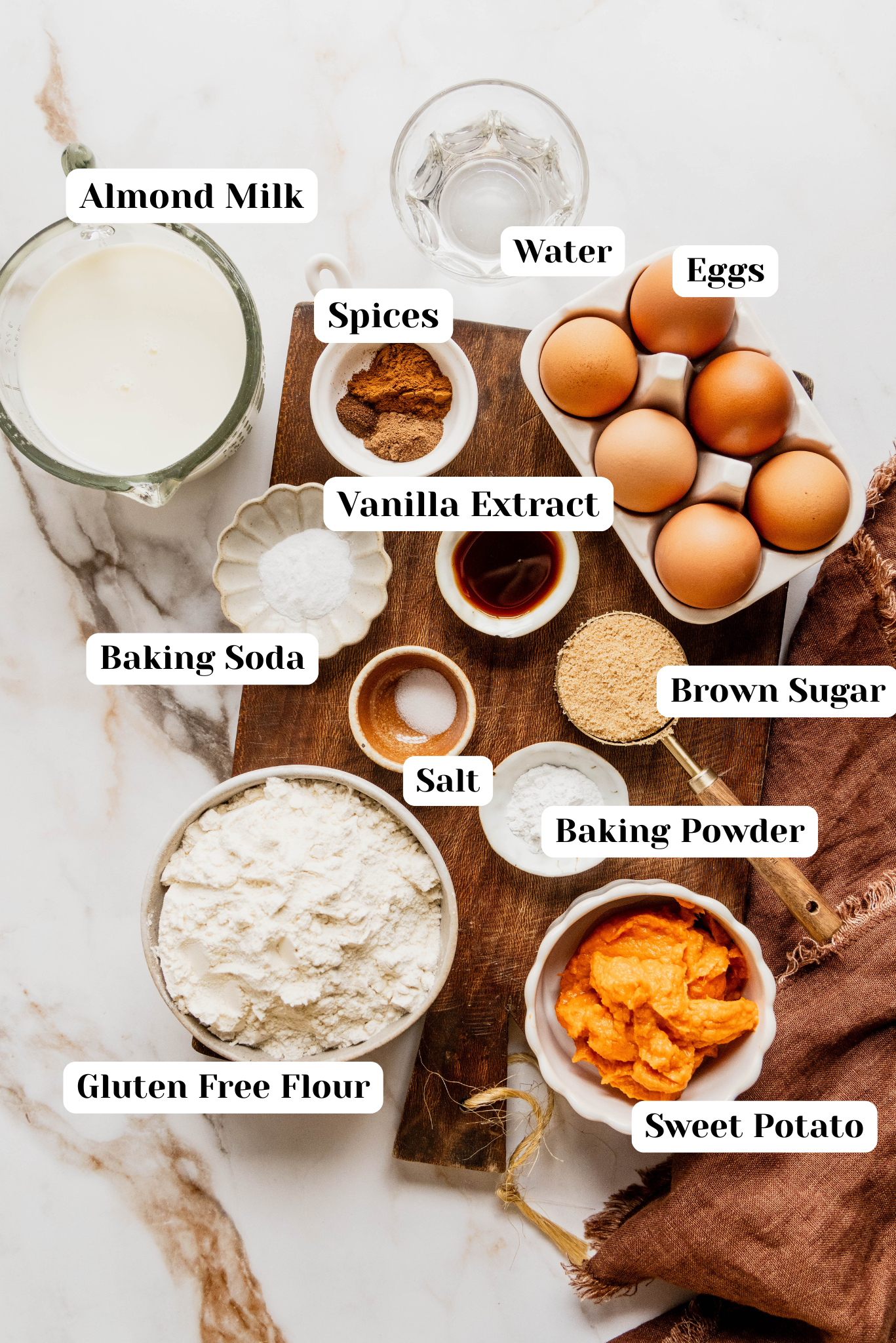 Overhead ingredient shot for gluten free sweet potato pancakes featuring gluten free flour, sweet potato puree, almond milk, eggs, brown sugar, baking powder, baking soda, salt, spices, vanilla extract, and water arranged on a wooden board over a white marble background.