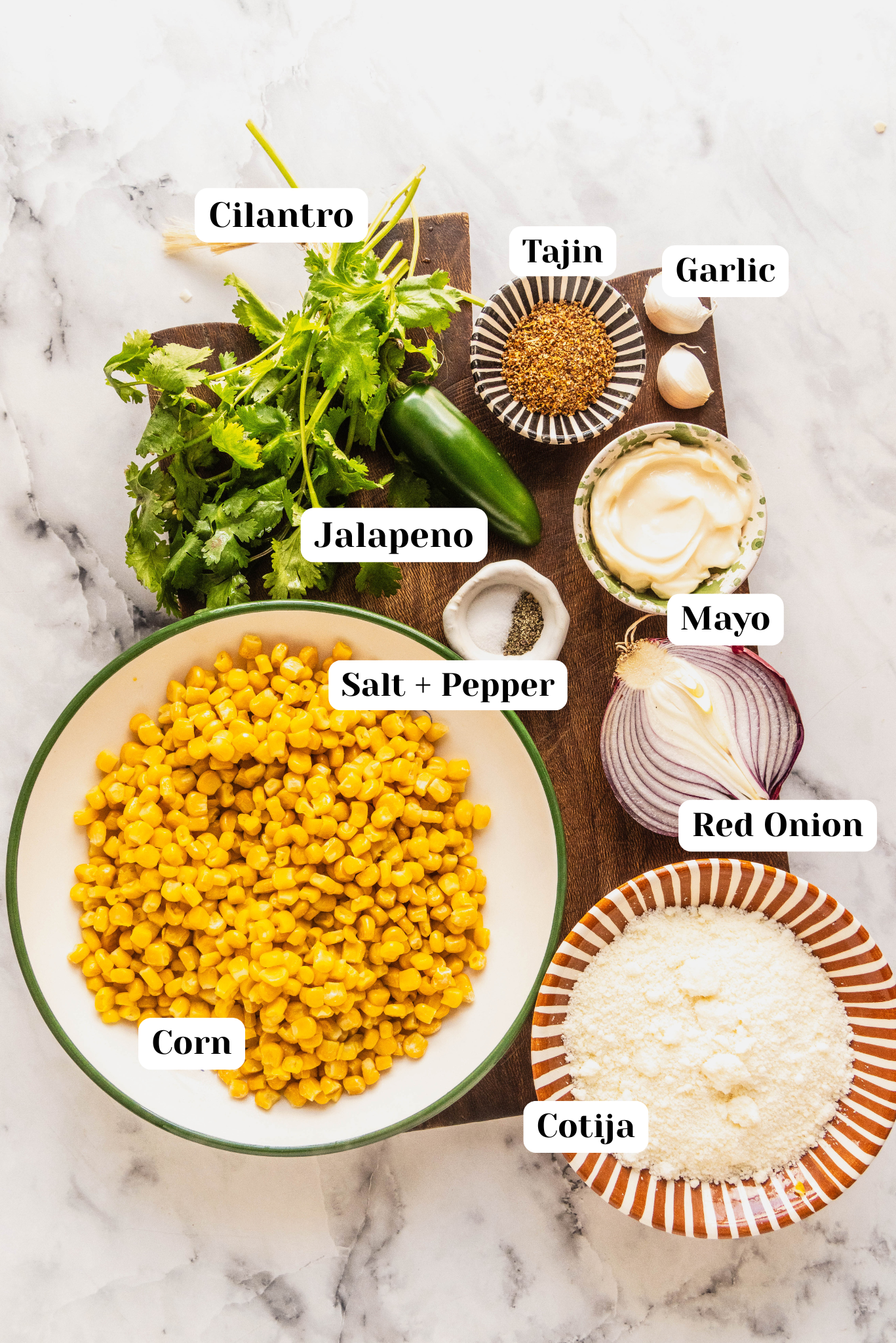 Overhead ingredient shot for Elotes corn dip featuring a bowl of sweet corn, crumbled cotija cheese, mayo, jalapeño, fresh cilantro, red onion, garlic, Tajín seasoning, and salt and pepper arranged on a wood board over a white marble surface.
