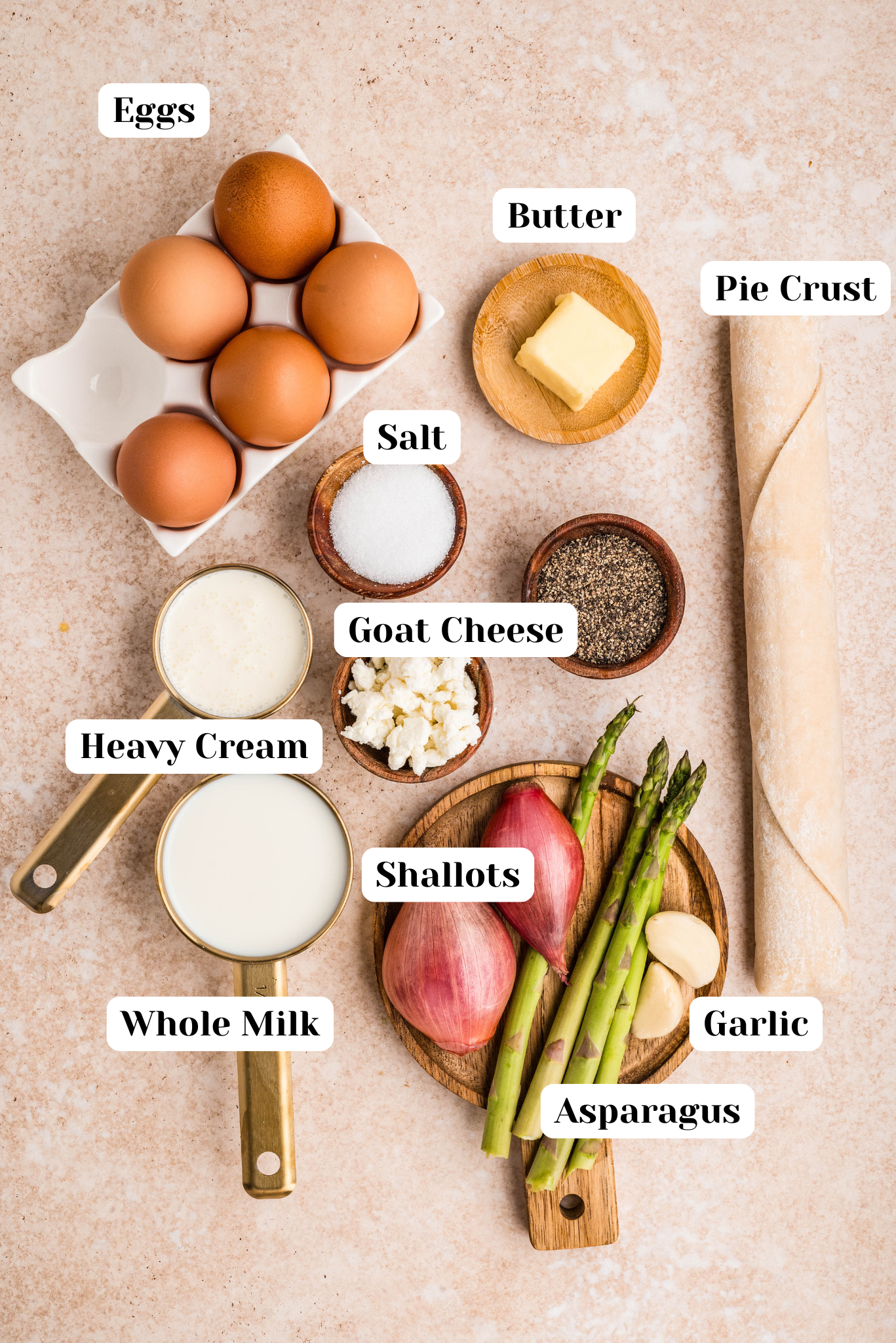 Overhead view of labeled ingredients for asparagus and goat cheese quiche including eggs, pie crust, butter, goat cheese, whole milk, heavy cream, asparagus, shallots, garlic, salt, and pepper arranged on a neutral background.