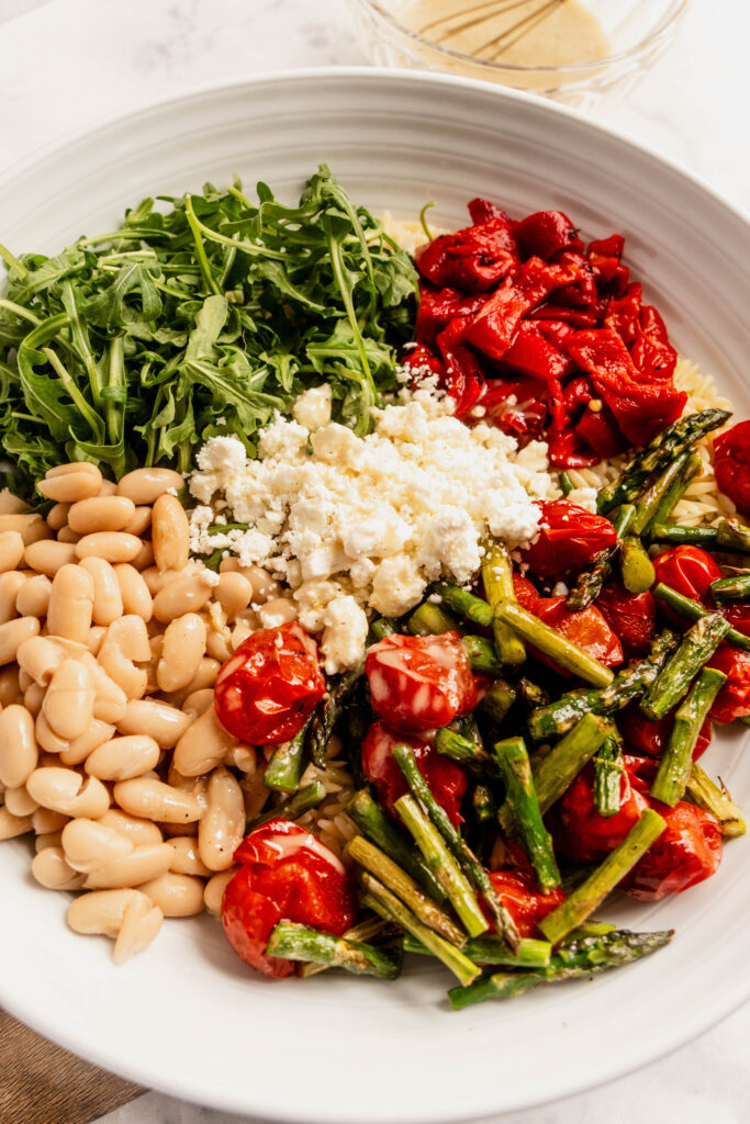 “Close-up of a plated asparagus orzo salad with roasted asparagus, blistered cherry tomatoes, white beans, arugula, roasted red peppers, crumbled feta, and lemon Dijon vinaigrette in a white bowl.”
