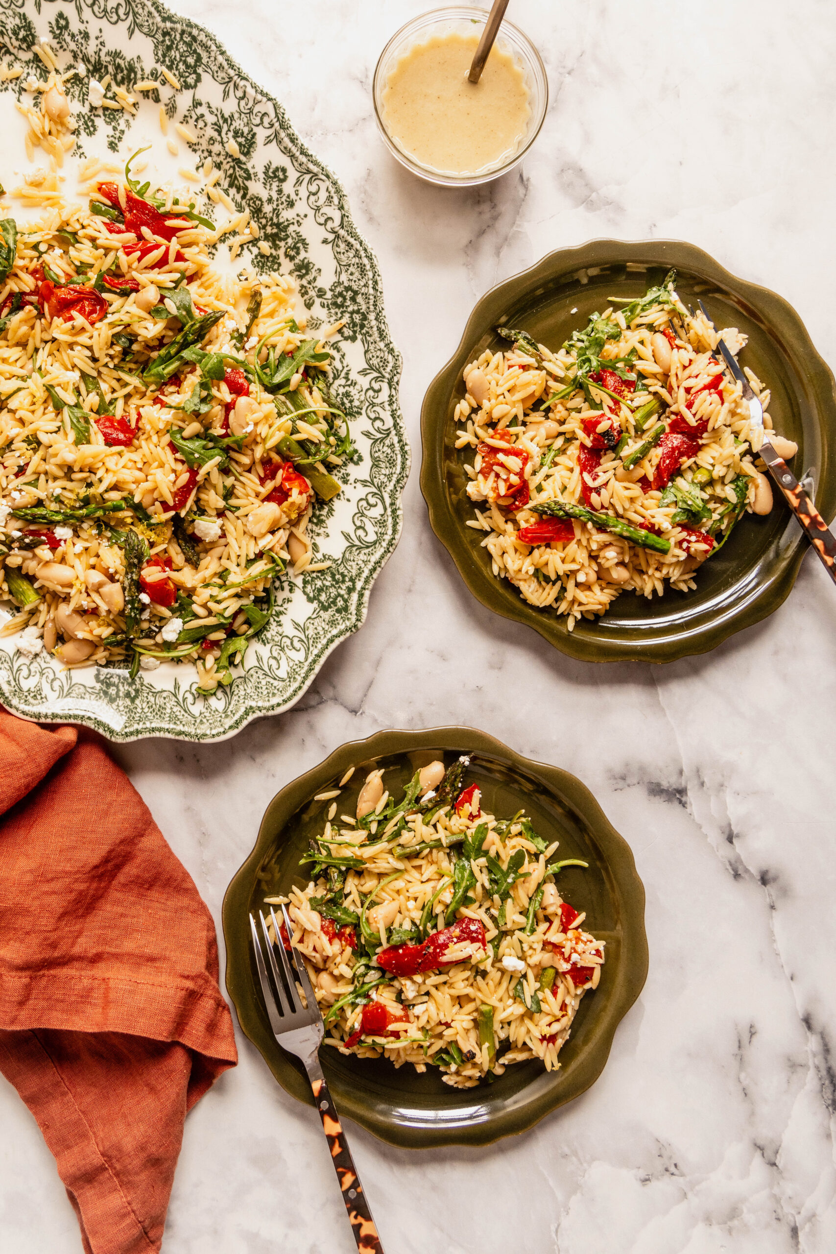 “Overhead view of asparagus orzo salad served family-style and in individual bowls, featuring roasted asparagus, cherry tomatoes, white beans, arugula, feta, and lemon Dijon vinaigrette on a white marble table.”