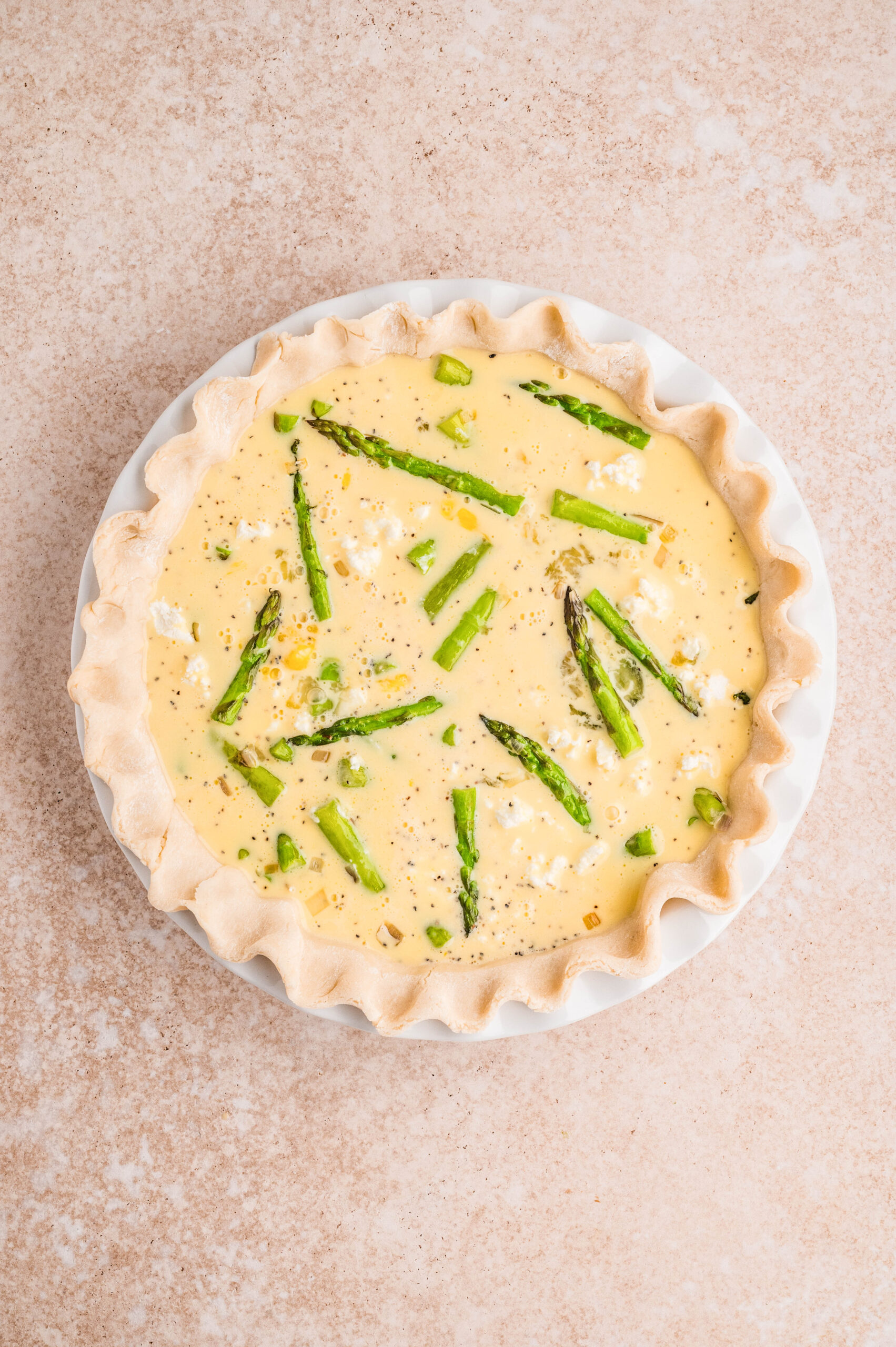 Overhead view of an unbaked asparagus and goat cheese quiche in a white pie dish, showing fresh asparagus pieces and crumbled goat cheese suspended in a creamy egg mixture inside a raw flaky crust on a neutral background.