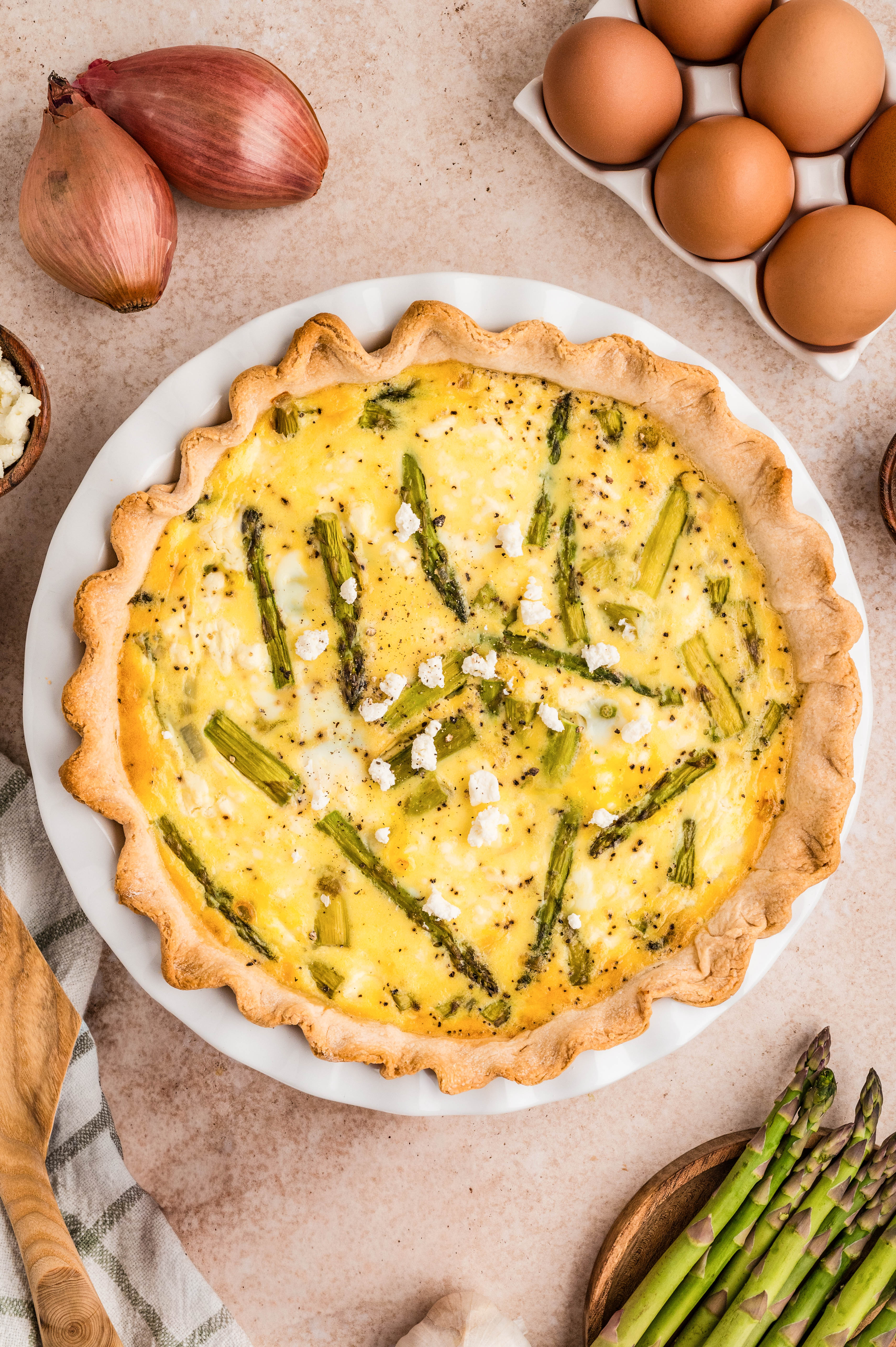 Overhead view of a whole asparagus and goat cheese quiche in a white pie dish, featuring a flaky golden crust and creamy egg filling dotted with tender asparagus and crumbled goat cheese, styled with fresh asparagus, shallots, eggs, and a wooden spatula on a neutral background.