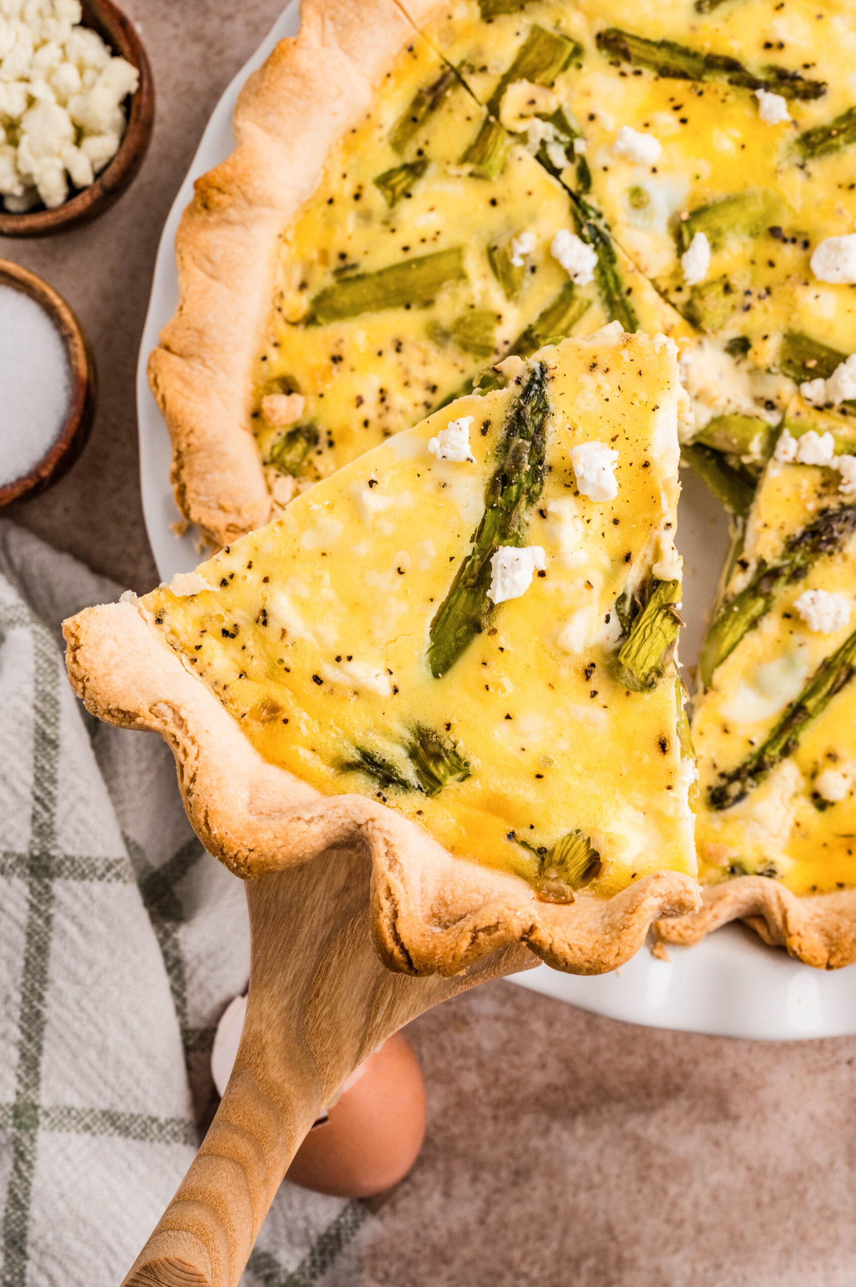 Overhead view of a slice of asparagus and goat cheese quiche lifted on a wooden spatula, showing tender asparagus pieces and creamy goat cheese baked into a golden egg filling and flaky crust, with the whole quiche, eggs, and fresh asparagus styled on a neutral background.