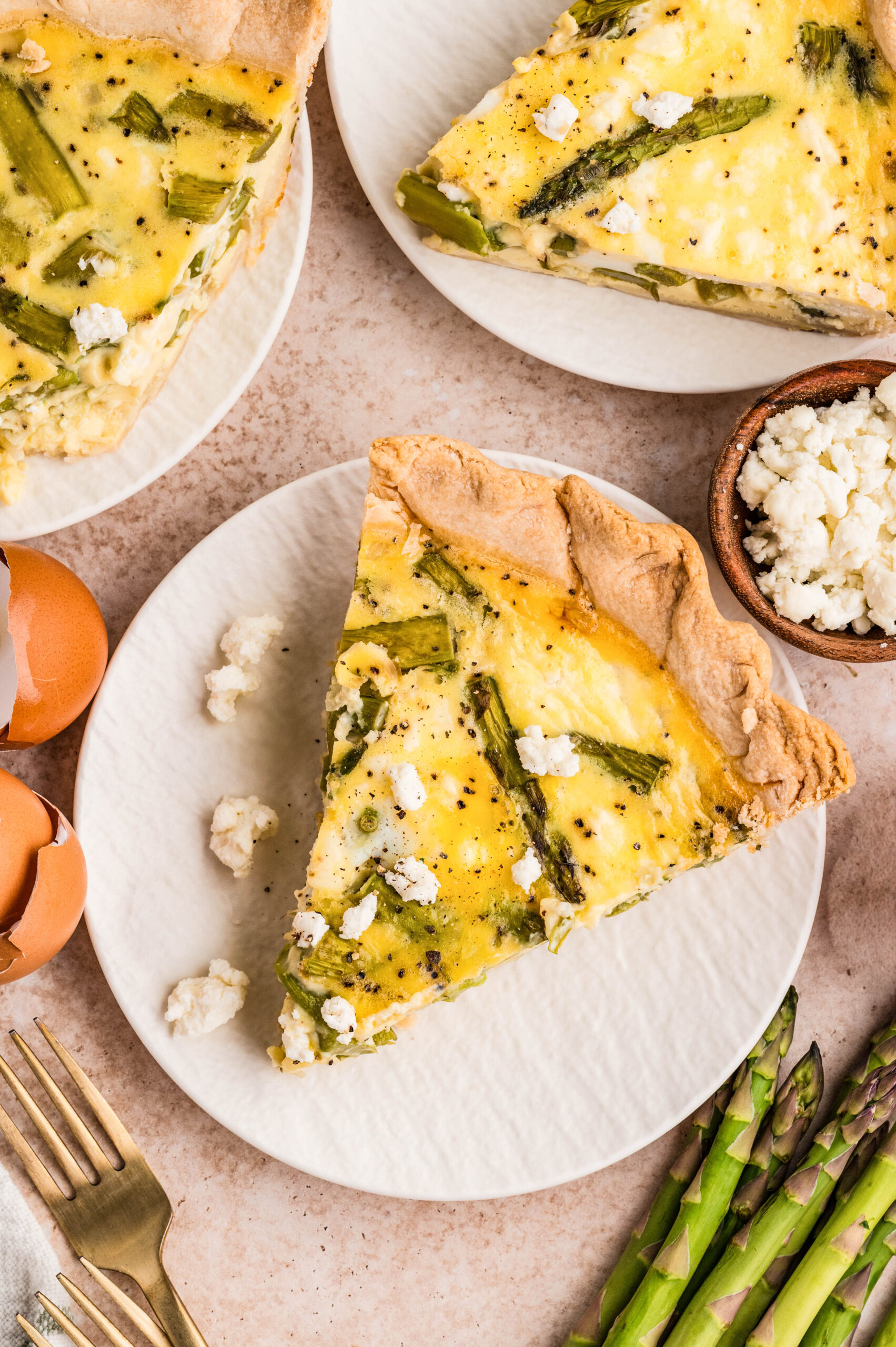 Overhead close-up of a slice of asparagus and goat cheese quiche on a white plate, showing tender asparagus spears and creamy goat cheese in a flaky golden crust, with whole quiche, fresh asparagus, eggs, and shallots styled on a neutral background.