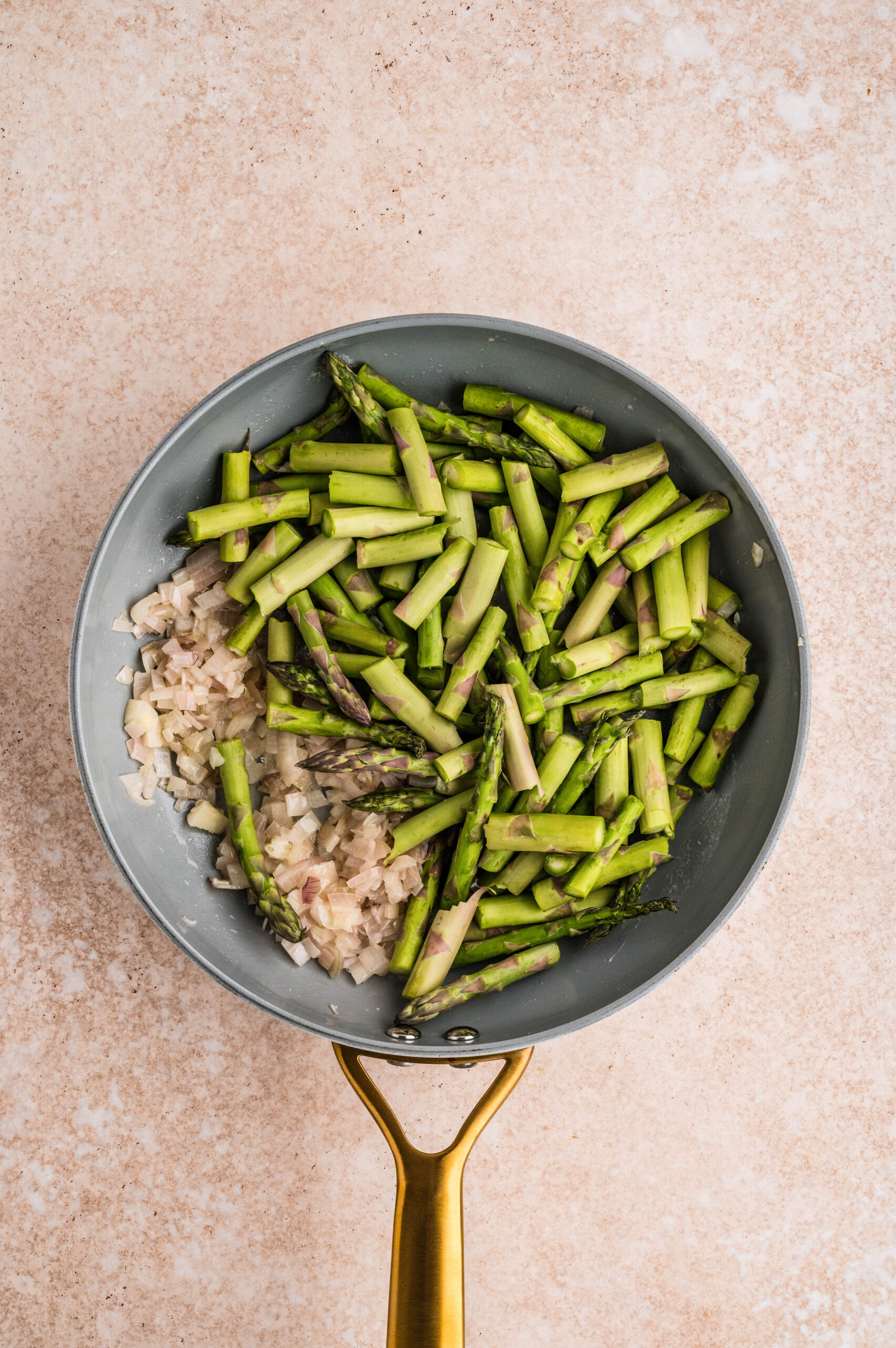 Overhead view of chopped asparagus and diced shallots sautéing in a skillet, ingredients prepared for an asparagus and goat cheese quiche, on a neutral background.