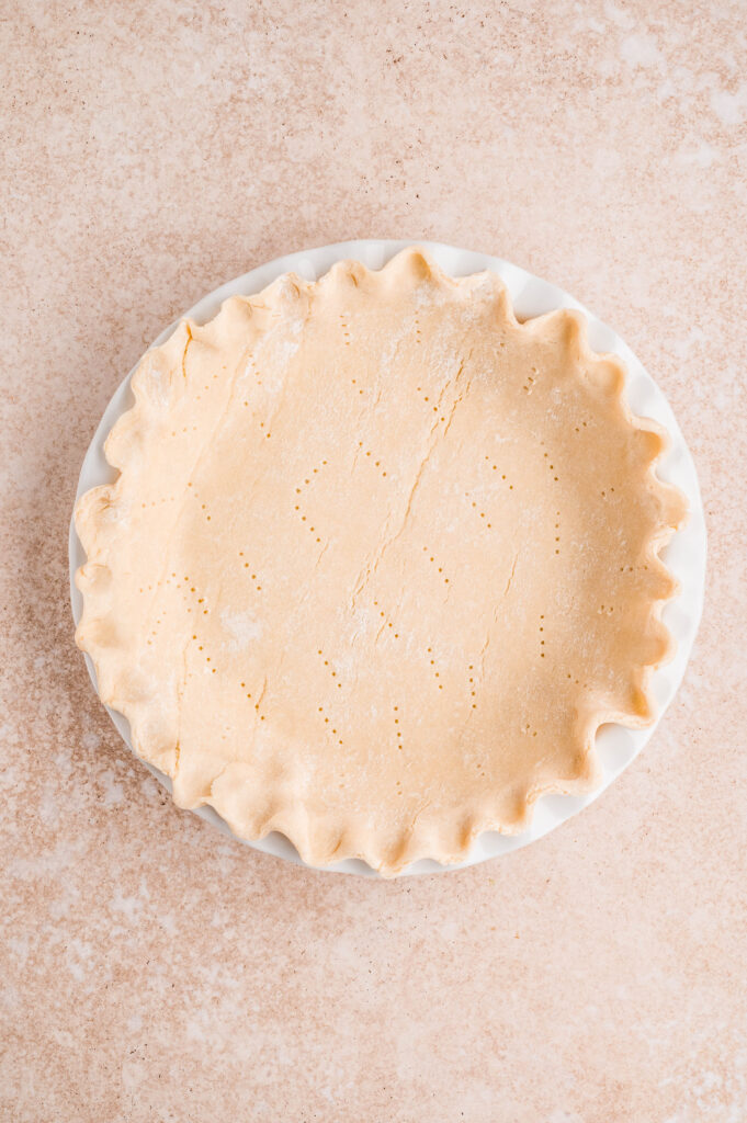 Overhead view of raw pie crust in a pie dish prepared for an asparagus and goat cheese quiche, on a neutral background.