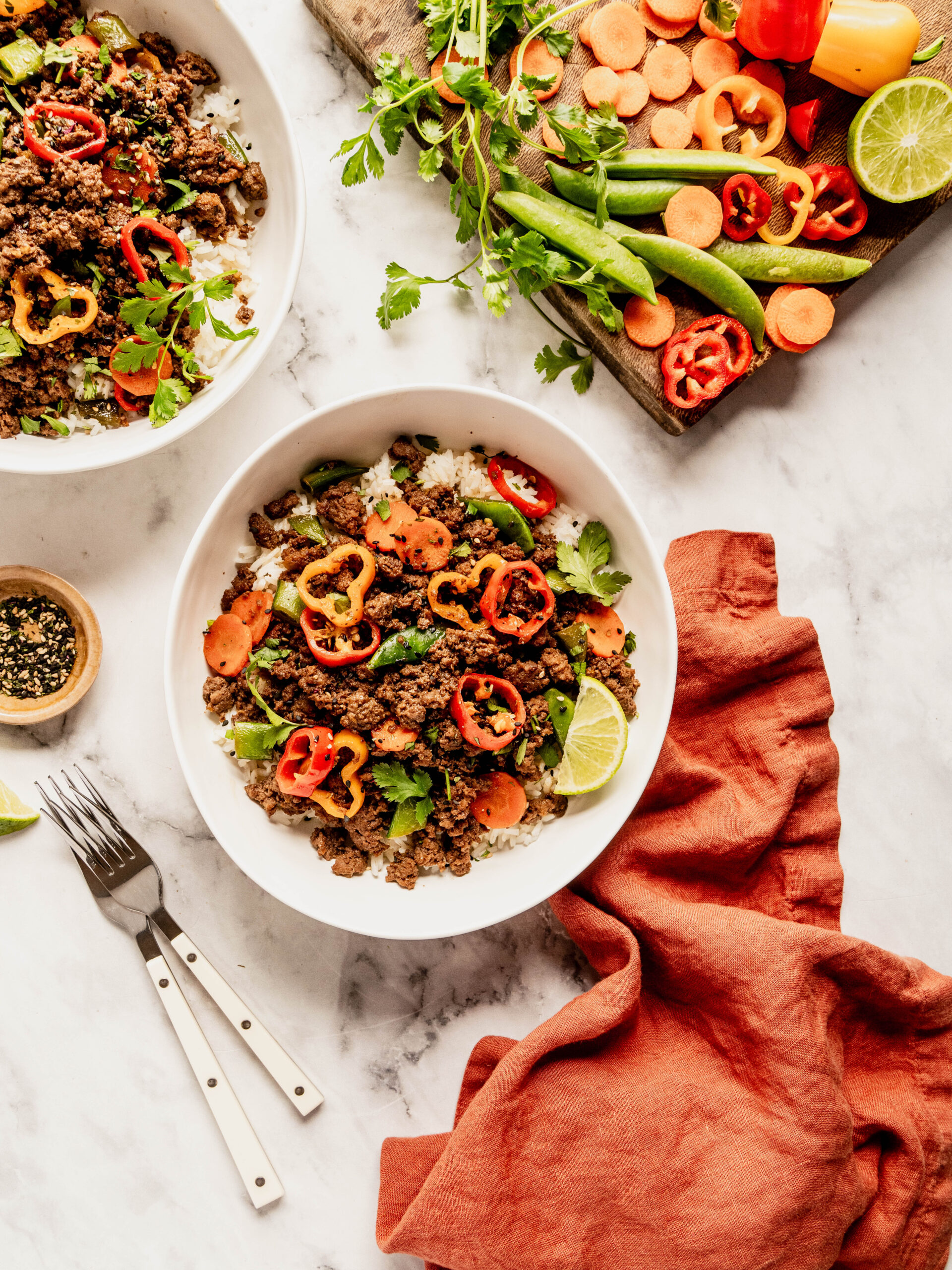 Warm, bright overhead photo of a Korean beef rice bowl served over white rice and topped with sautéed ground beef, sliced carrots, sugar snap peas, red and orange bell peppers, fresh cilantro, sesame seeds, and a lime wedge. Fresh vegetables and herbs are styled on a wooden board beside the bowl on a white marble background with a rust-colored linen napkin.