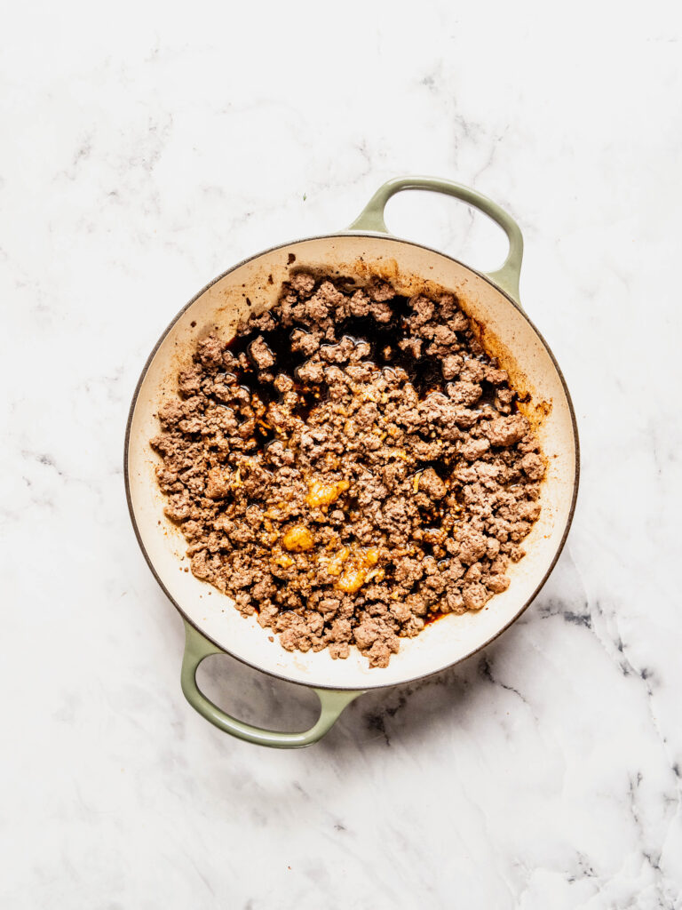 Overhead view of ground beef cooking in a skillet with soy sauce, honey, garlic, and ginger for a Korean beef rice bowl, showing the savory sauce pooling and caramelizing around the browned beef on a white marble background.