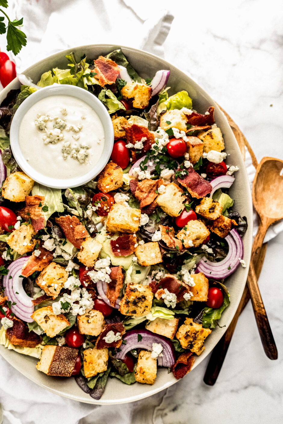 Chopped BLT salad in a large serving bowl topped with crispy bacon pieces, golden croutons, crumbled blue cheese, grape tomatoes, and sliced red onion over chopped lettuce, served with a small bowl of creamy blue cheese dressing on a marble surface with fresh herbs nearby.