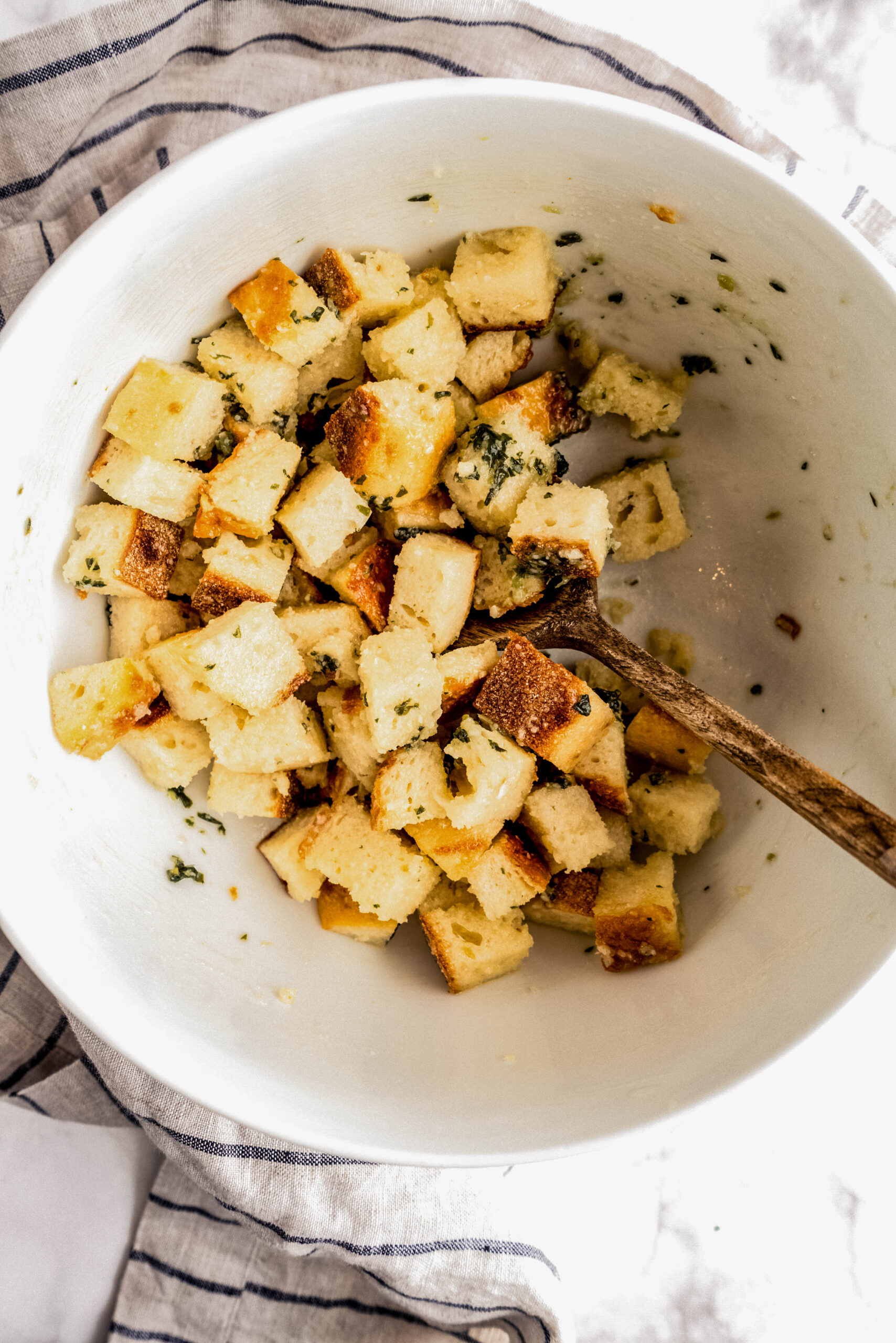 Overhead view of homemade Parmesan croutons in a large white mixing bowl, showing golden toasted bread cubes coated with olive oil, grated Parmesan, and fresh herbs, with a wooden spoon and striped kitchen towel on a marble countertop.