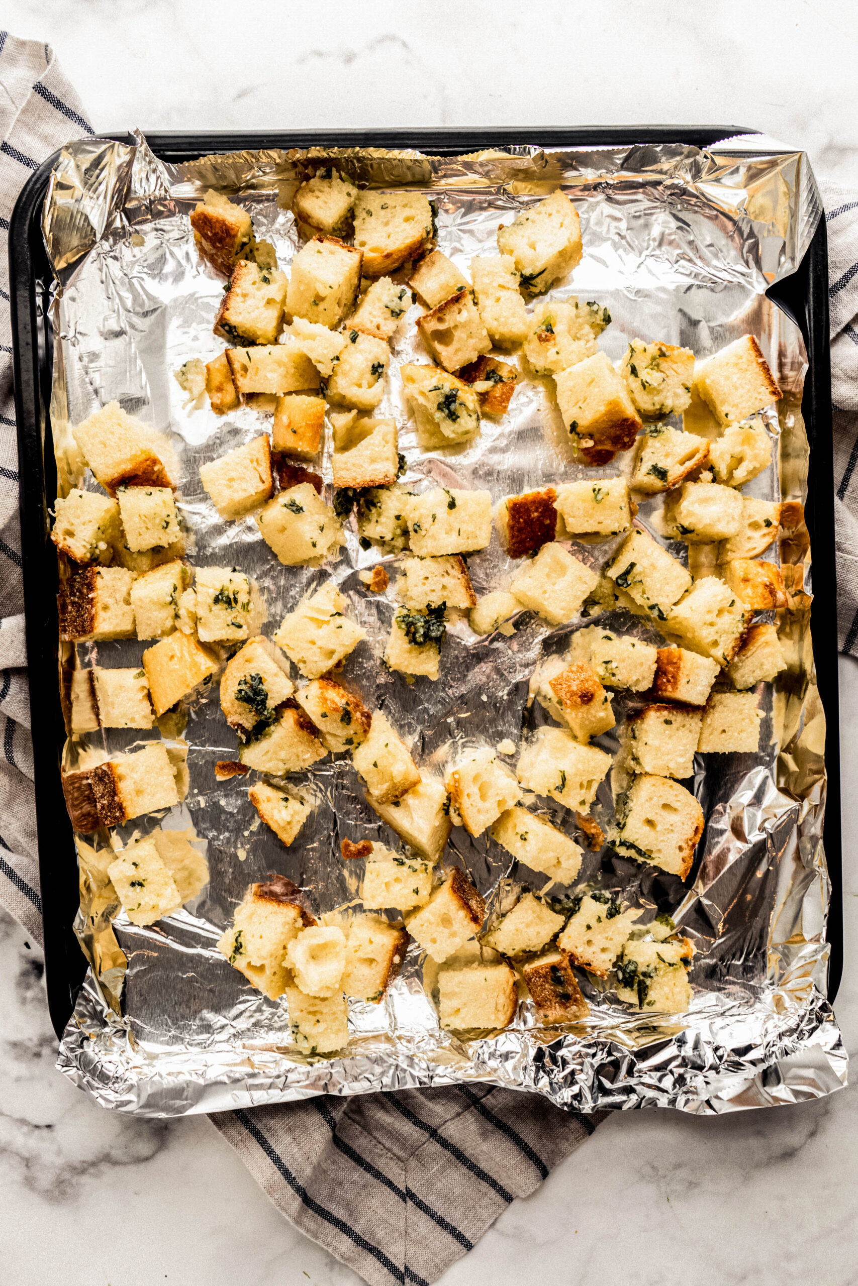 Overhead view of homemade Parmesan croutons baked on a foil-lined sheet pan, showing golden brown bread cubes coated in olive oil, grated Parmesan, and fresh herbs on a marble countertop with a neutral kitchen towel underneath.
