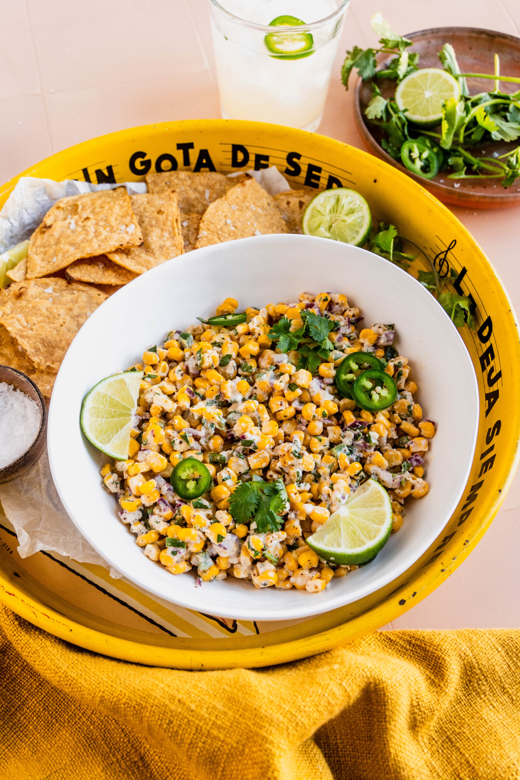 Up close view of Elotes corn dip served in a white bowl, topped with sliced jalapeños, fresh cilantro, and lime wedges, surrounded by tortilla chips on a bright yellow tray with a small bowl of flaky salt and a fresh lime drink nearby.