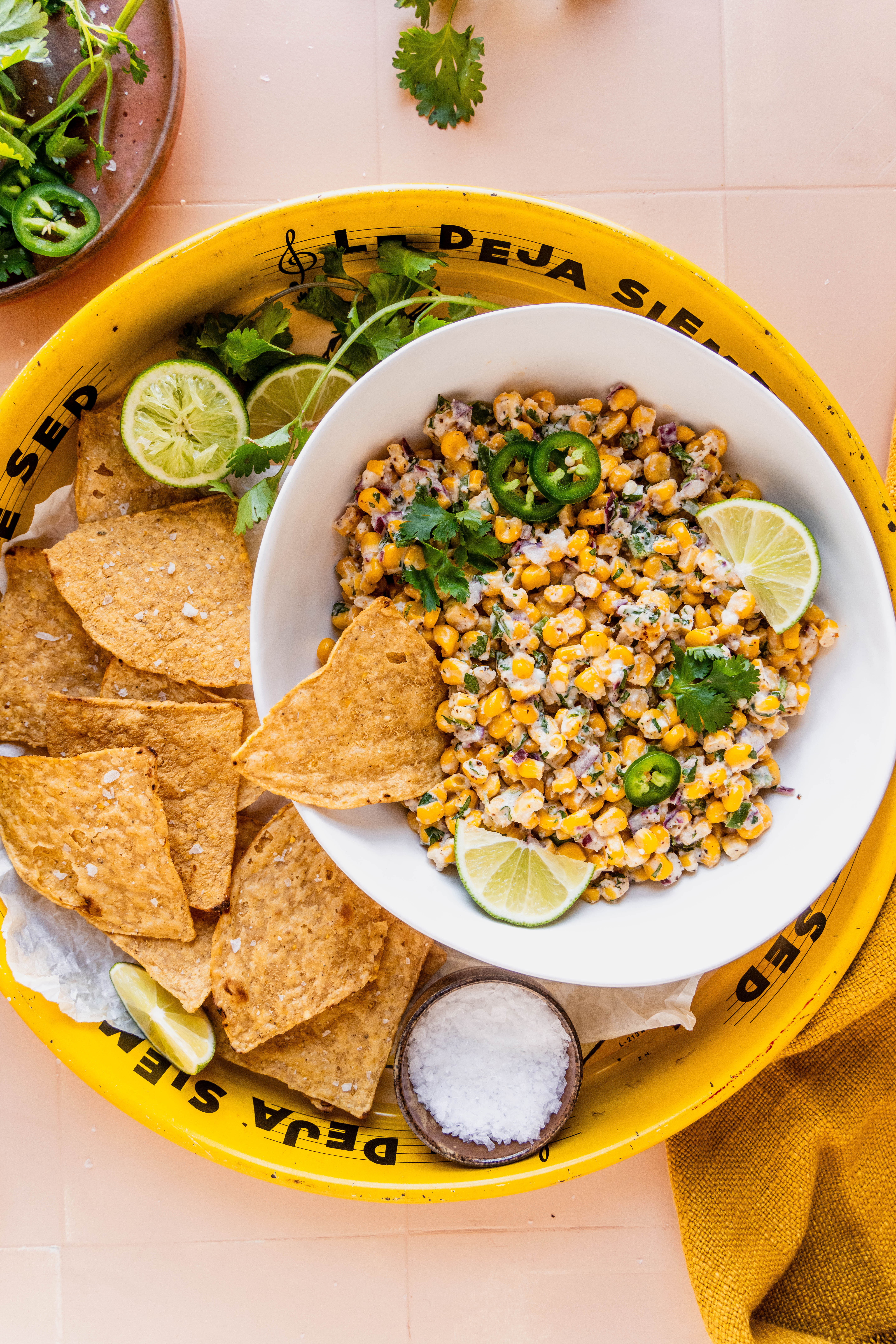 Overhead view of Elotes corn dip served in a white bowl, topped with sliced jalapeños, fresh cilantro, and lime wedges, surrounded by tortilla chips on a bright yellow tray with a small bowl of flaky salt and a fresh lime drink nearby.