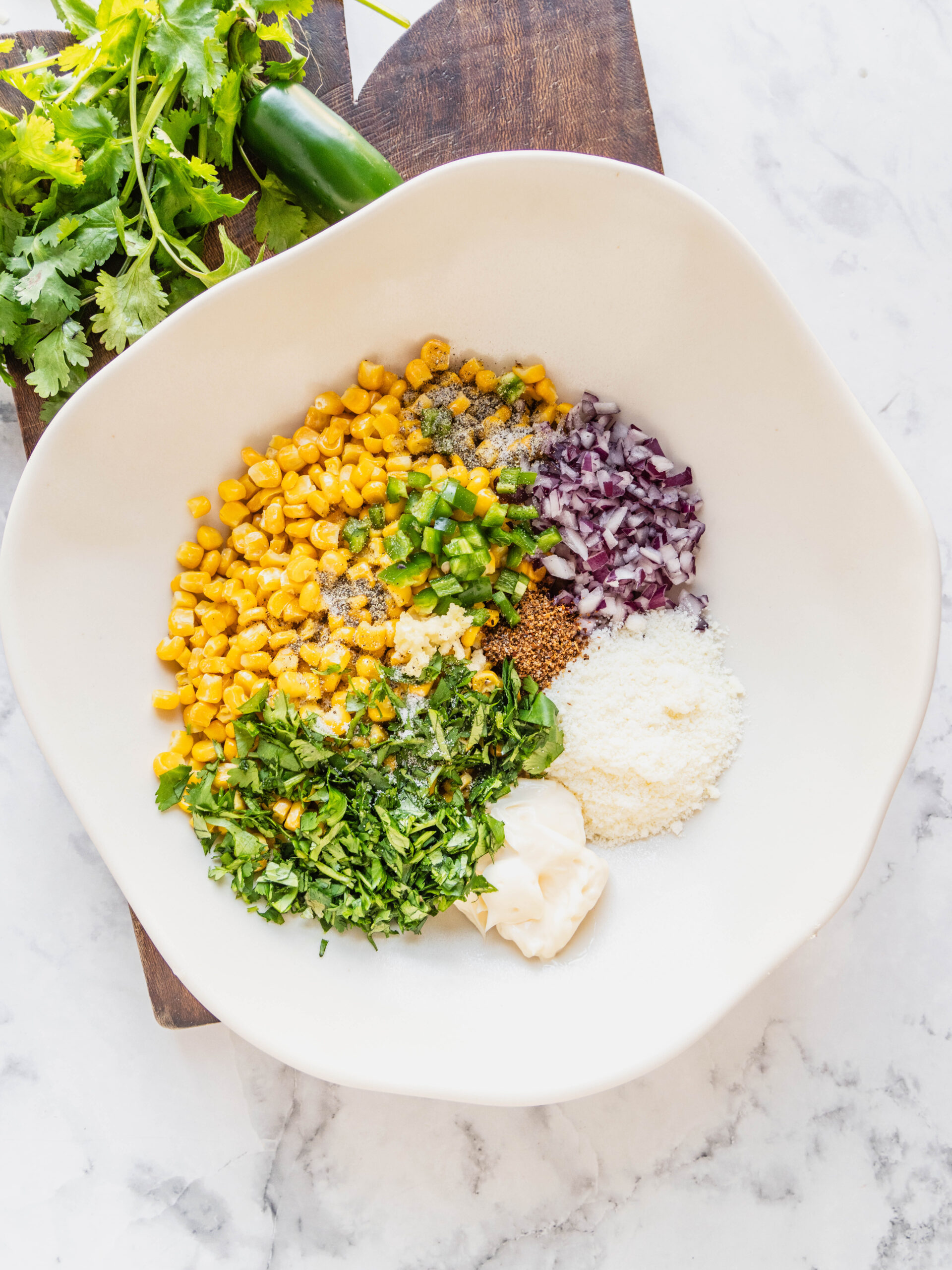 Overhead view of ingredients in a white bowl for Elotes corn dip, including sweet corn, chopped jalapeño, red onion, garlic, cilantro, cotija cheese, mayo, Tajín, and salt and pepper, arranged before mixing on a marble surface.