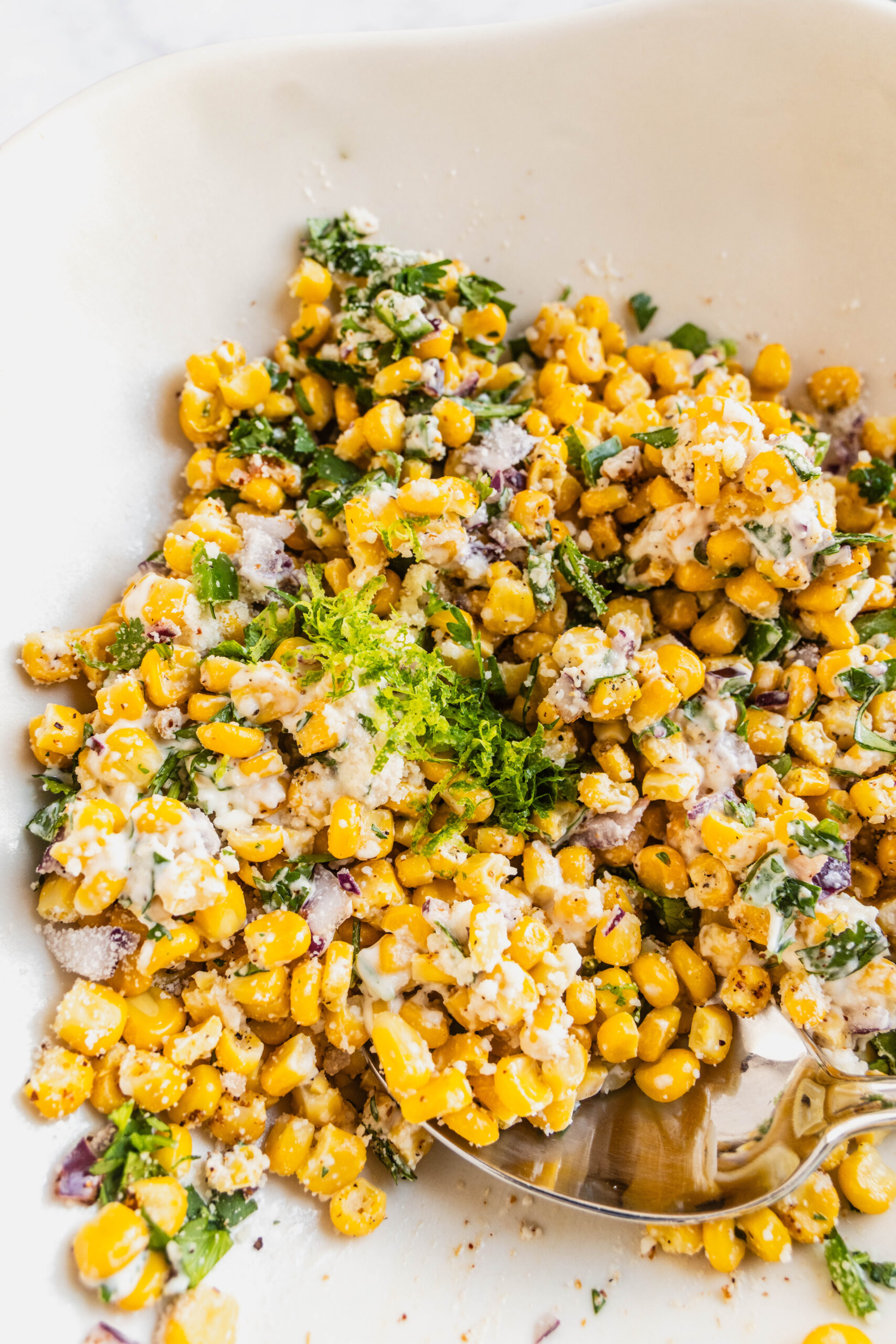 Close-up of Elotes corn dip in a white bowl, featuring sweet yellow corn tossed with creamy mayo, cotija cheese, chopped cilantro, red onion, jalapeño, lime zest, and Tajín seasoning, with a spoon scooping from the side.