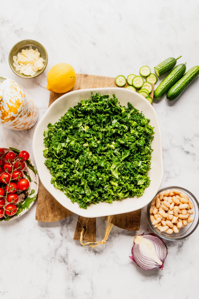 Overhead view of chopped kale in a white bowl for kale and cannellini bean salad, surrounded by grape tomatoes, Persian cucumbers, cannellini beans, red onion, lemon, olive oil, and shaved Pecorino on a marble countertop.